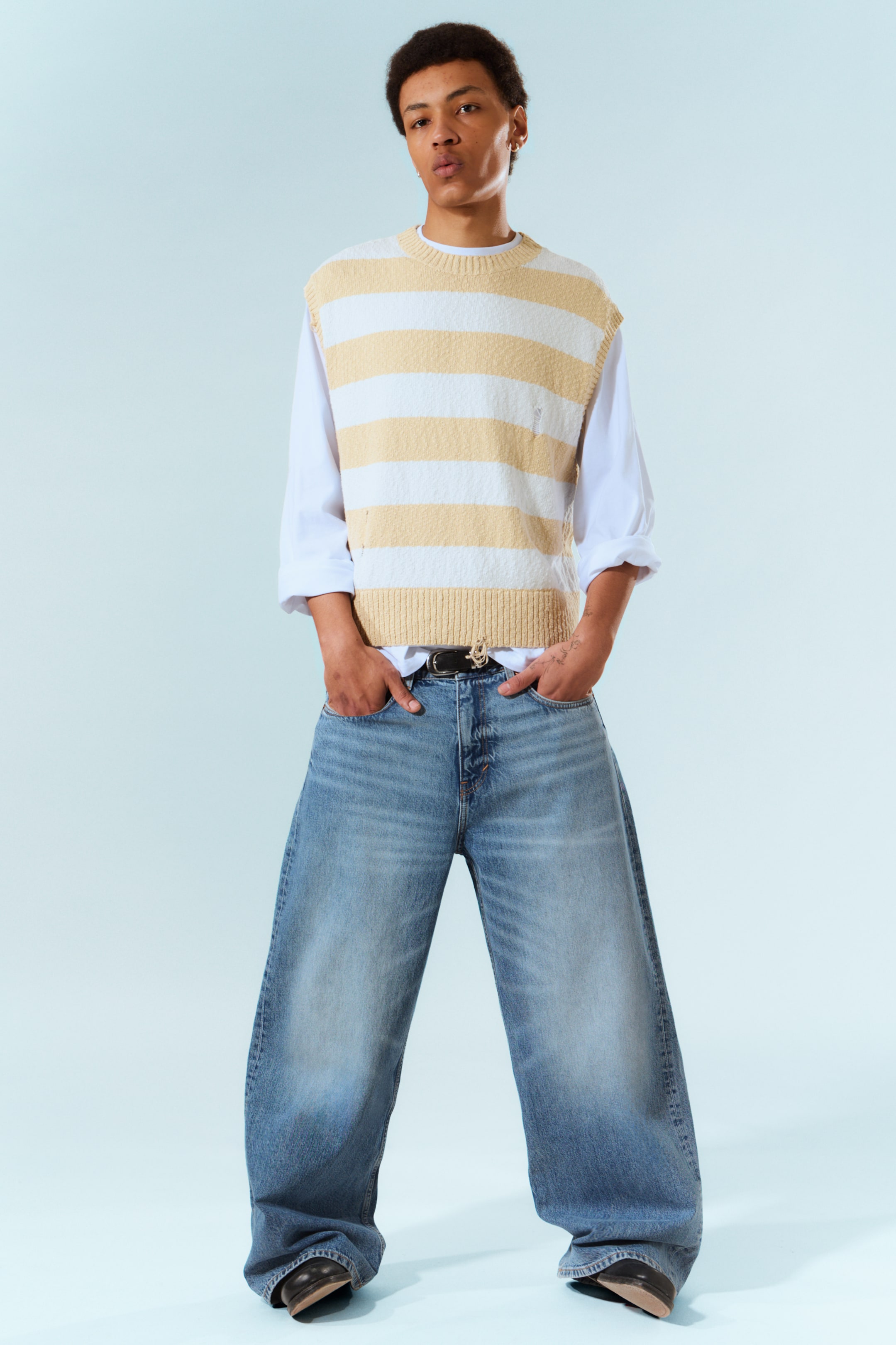 Ver imagem maior: Um colete de malha texturizada com riscas horizontais em creme e branco, com detalhes desgastados e uma gola redonda, está a ser usado sobre uma camisa branca de mangas compridas. Calças de ganga largas e de lavagem clara são usadas com um cinto preto, que exibe uma fivela em tom dourado.