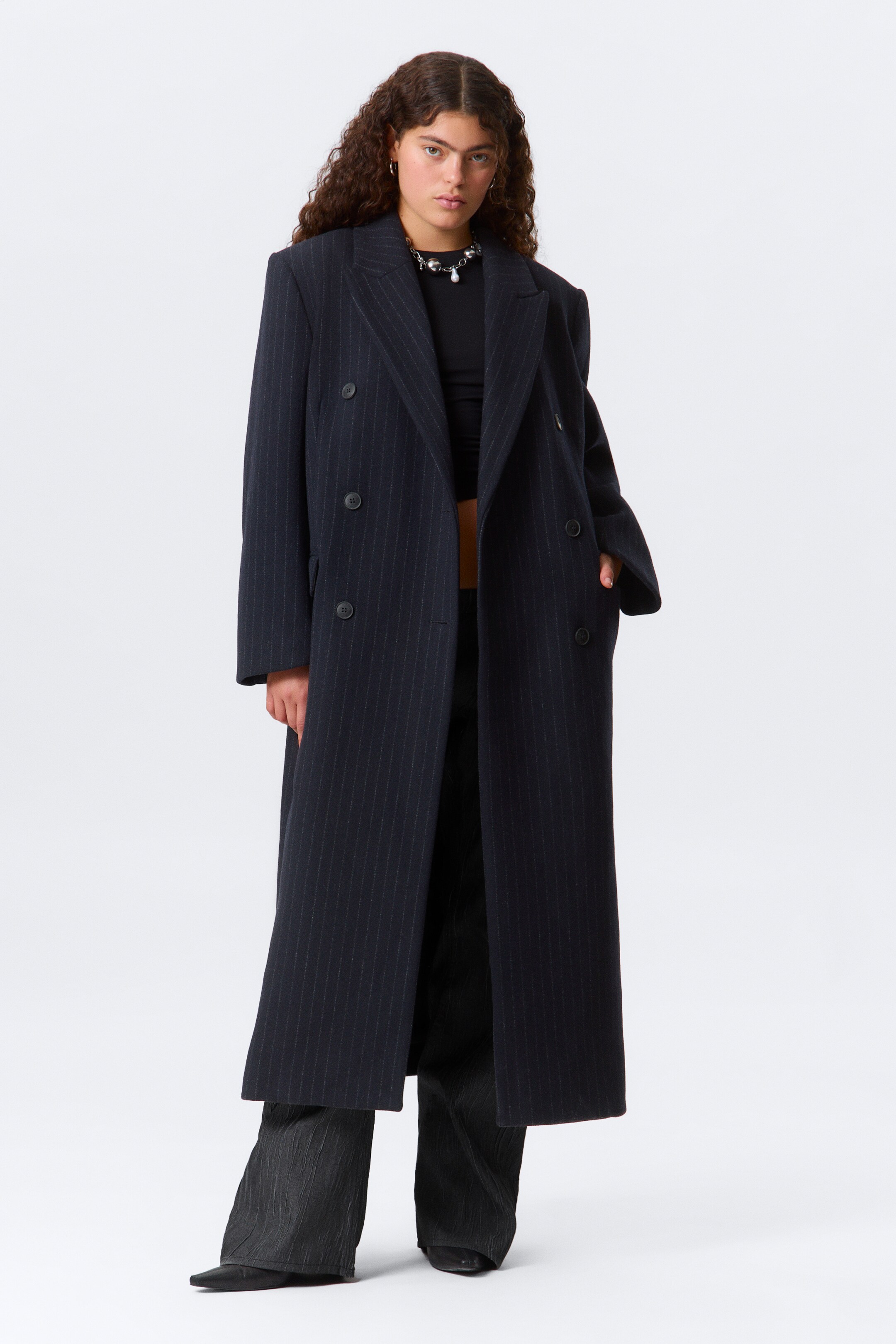 Vis større bilde: En mørk dobbelkneppet frakk med nålestriper blir båret forovervendt, med brede hakkslag og strukturerte skuldre. Den har full lengde som strekker seg til anklene, med to rader med mørke knapper foran og knapper på mansjettene, og den er lagd over mørke bukser.
