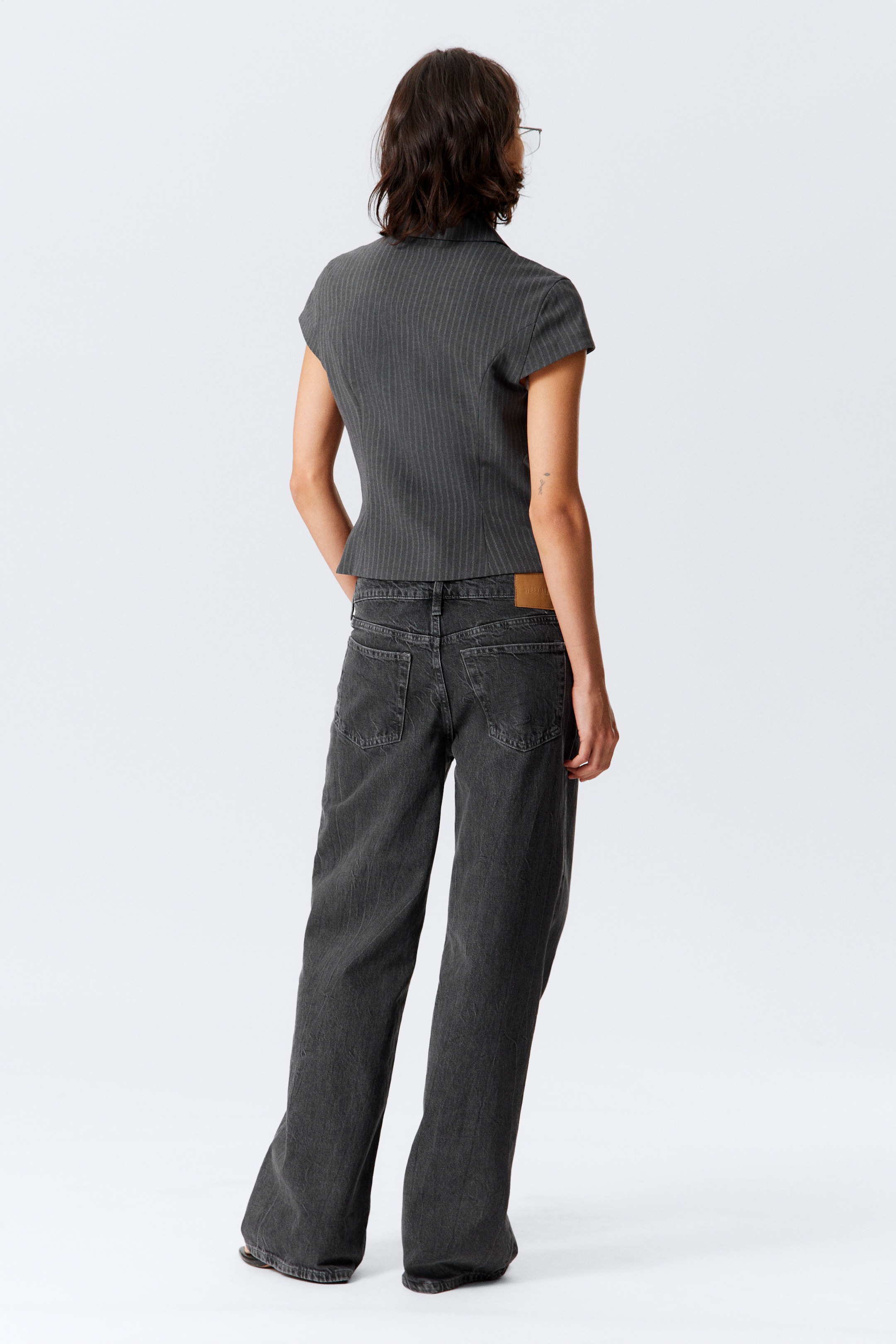 Grotere afbeelding bekijken: Een donkergrijs gestreept, kort mouwloos topje, met een aansluitende taille, wordt van de achterkant gepresenteerd. Het wordt gestyled met donkergrijze, wijde jeans, die twee achterzakken en een lichtbruine patch aan de rechterkant van de tailleband tonen.