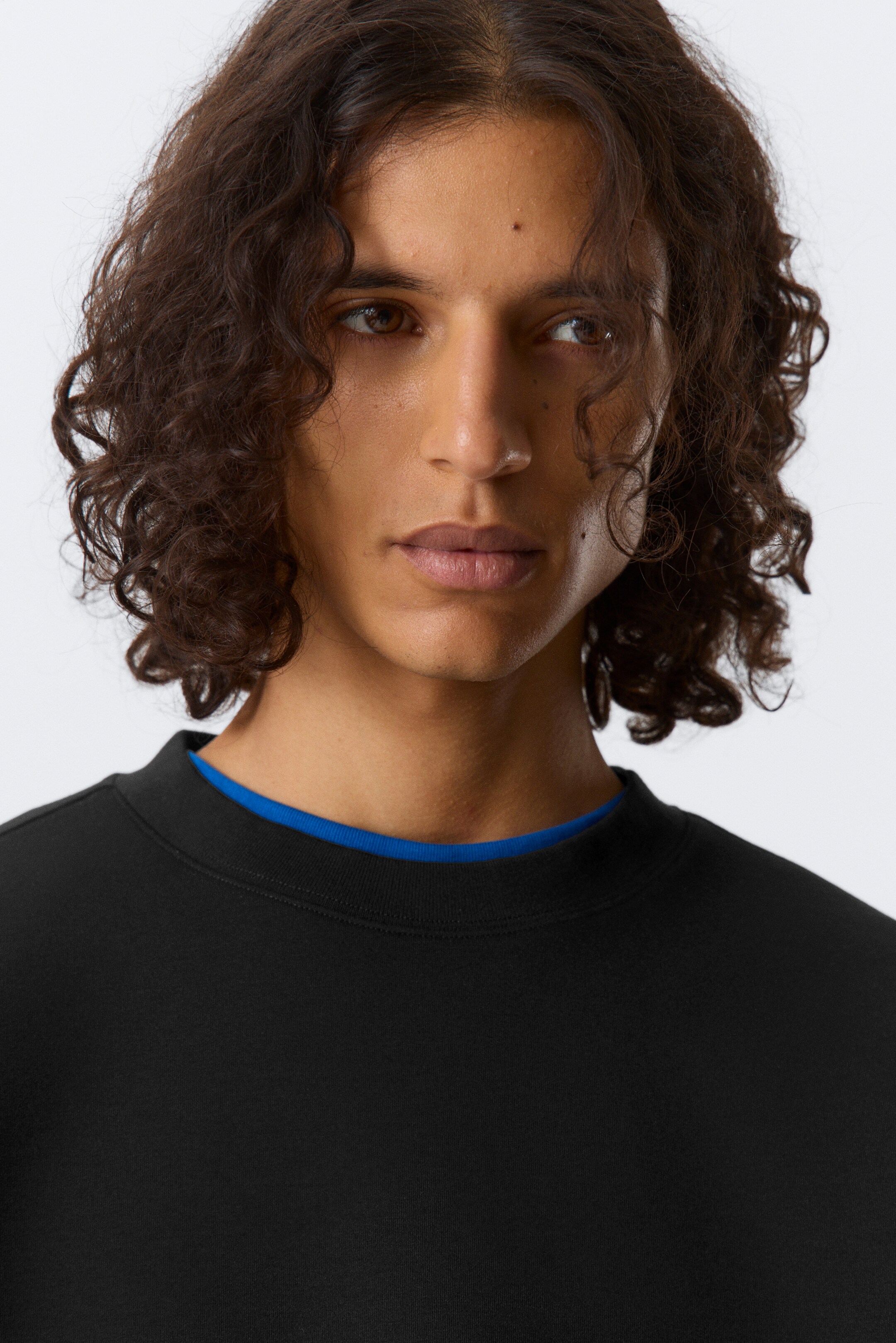 Se større billede: En sort crewneck sweatshirt vises fra en let vinklet frontvisning. En blå beklædningsgenstand er synligt lagdelt under dens halsudskæring.