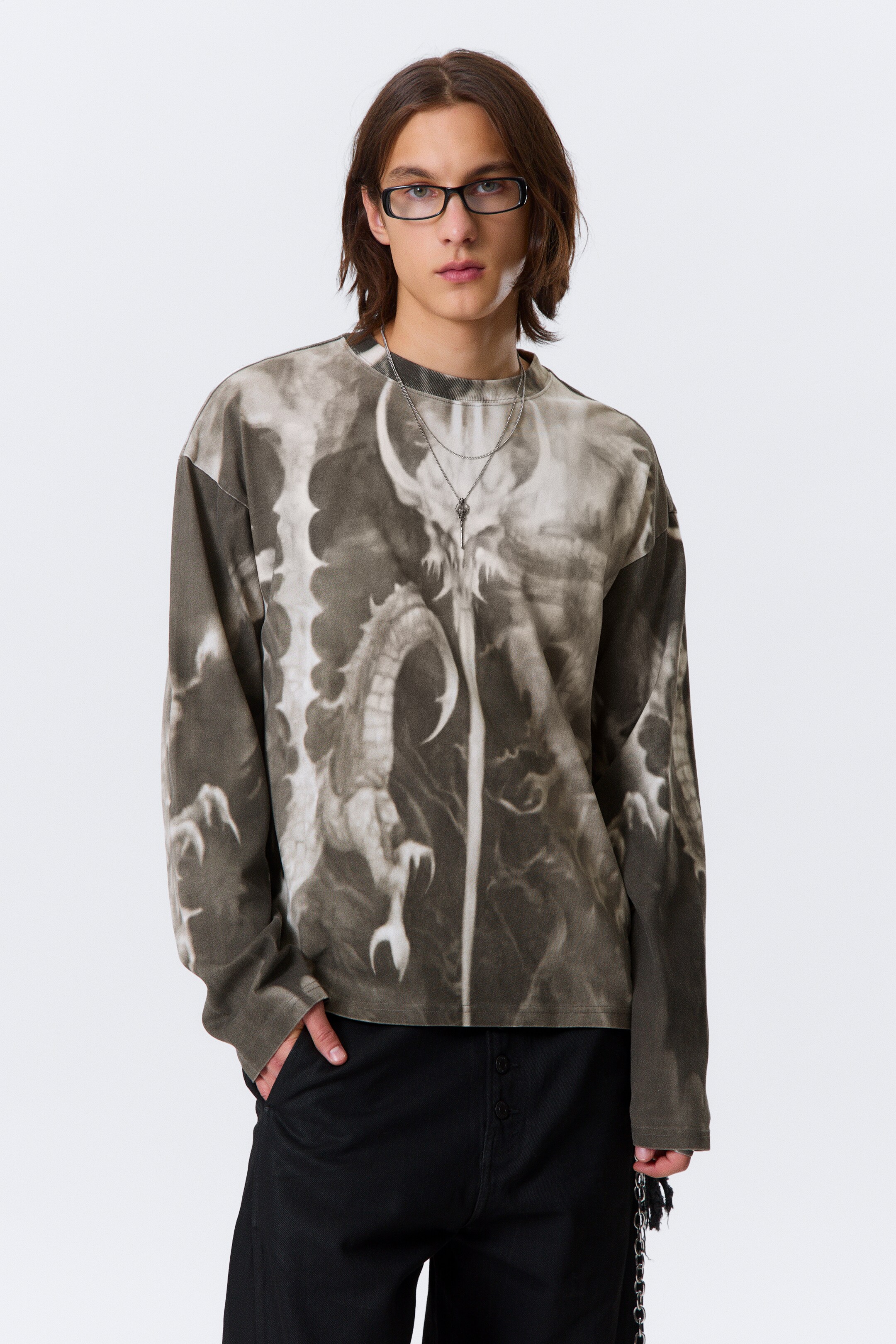 Agrandir l'image: Une chemise surdimensionnée à manches longues dans des tons de gris vieilli présente un imprimé graphique proéminent d'une créature mythique semblable à un dragon sur le devant. La chemise affiche un col rond et des manches amples, avec un collier en chaîne argentée superposé par-dessus.