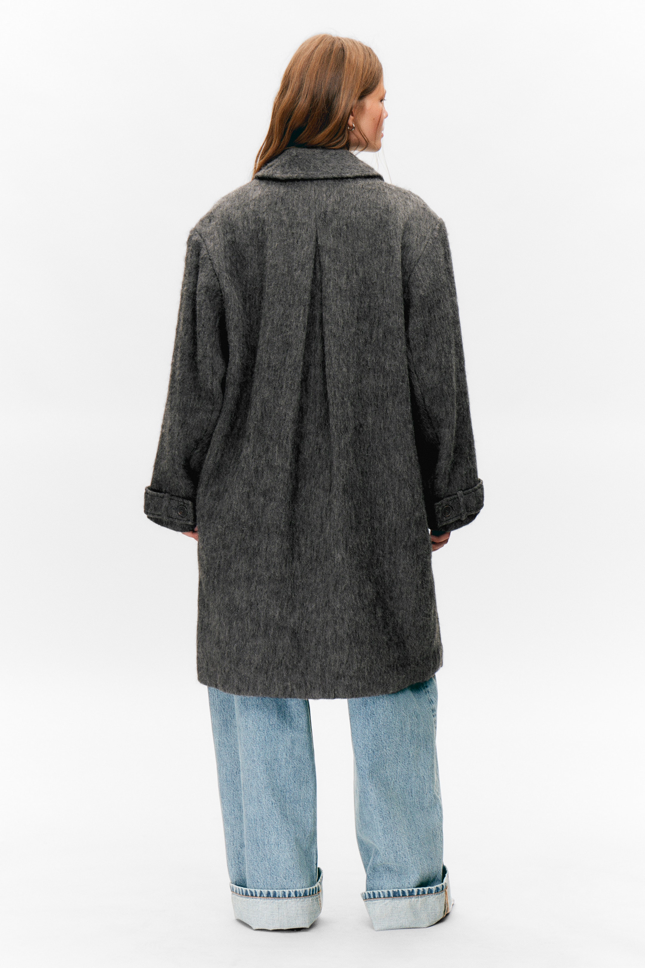 Ingrandisci l'immagine: Un cappotto grigio scuro viene visto da dietro, caratterizzato da un materiale testurizzato e peloso e da una vestibilità oversize e rilassata che si estende fino a metà coscia sopra jeans azzurri. Presenta maniche ampie con polsini arrotolati, uno dei quali mostra una linguetta con bottone, e una piega centrale corre lungo la schiena dal colletto.