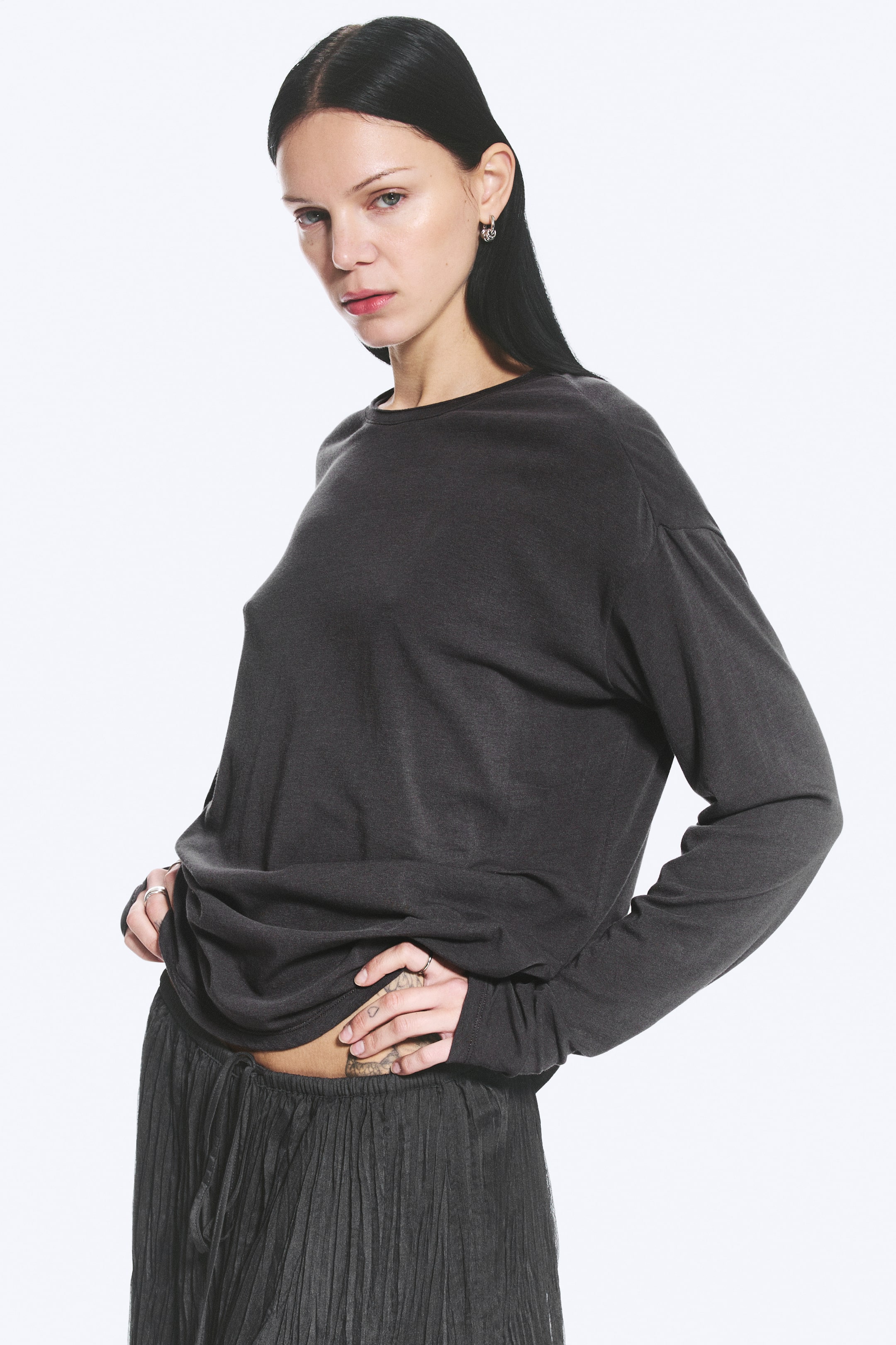 Visa större bild: Modellen bär en mörkgrå, lös, oversized, långärmad T-shirt med rund hals. T-shirten har en draperad känsla och modellen håller i fållen vid midjan och lyfter den något.