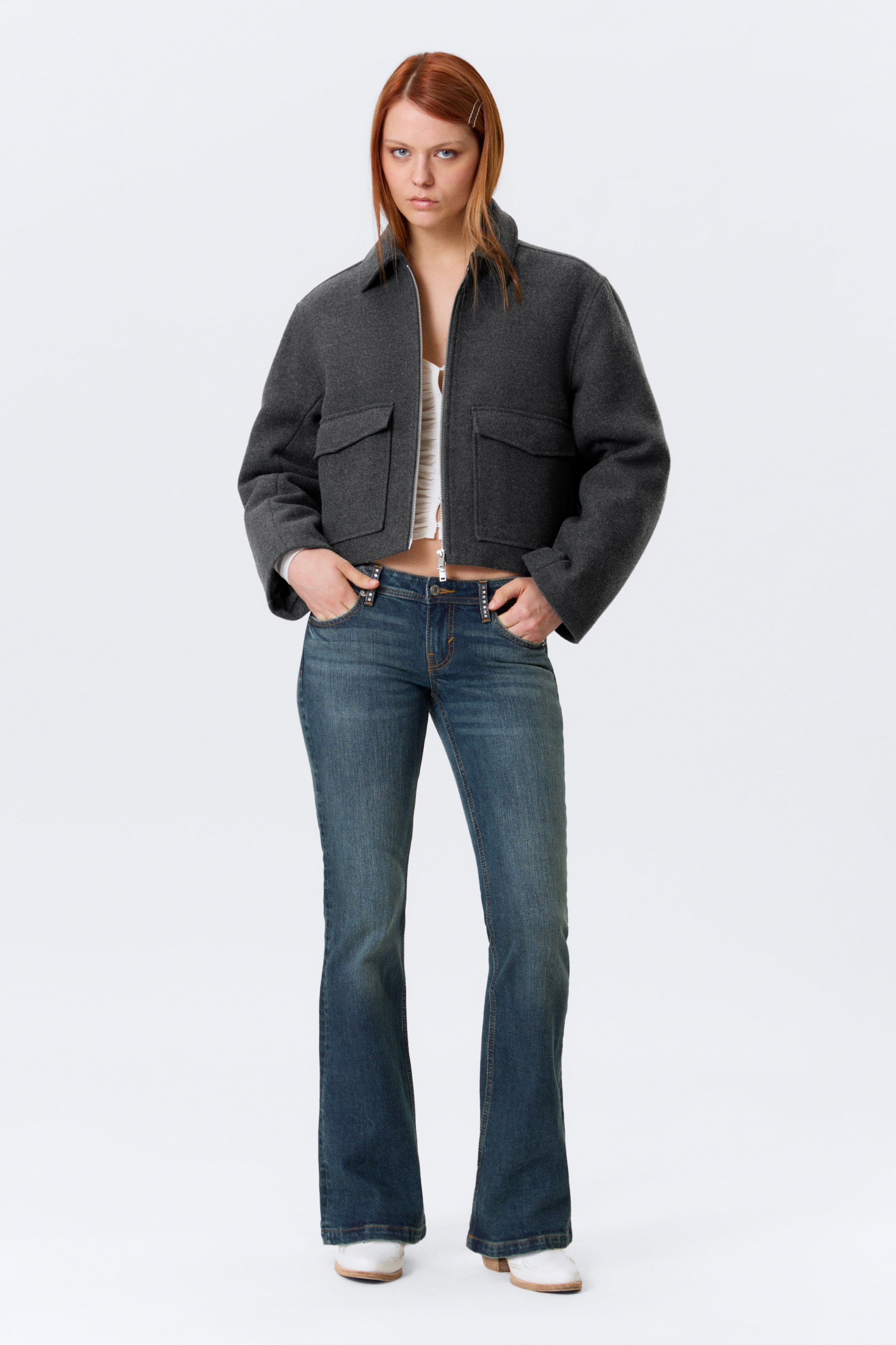 Grösseres Bild ansehen: Eine dunkelgraue, kurz geschnittene Jacke wird von vorne gezeigt, mit einem breiten Kragen, einer offenen Front, die ein weißes Top darunter enthüllt, zwei großen quadratischen Klappentaschen auf der Brust und weit geschnittenen Ärmeln.