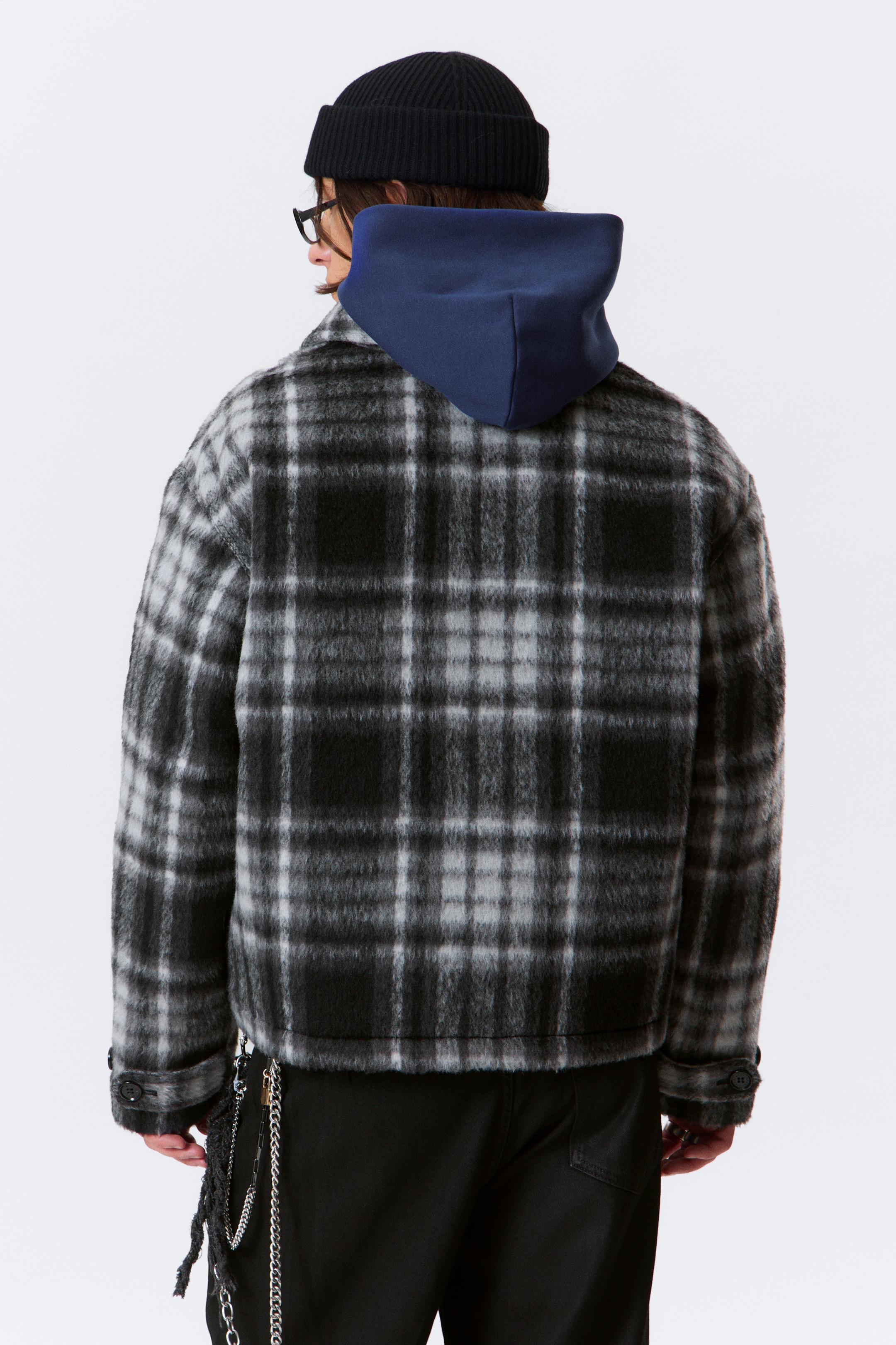 Visa större bild: En beskuren, svart, vit och grå rutig jacka med en luddig textur ses bakifrån. Den har knäppta manschetter och en rak fåll, medan en mörkblå hoodie ligger under.