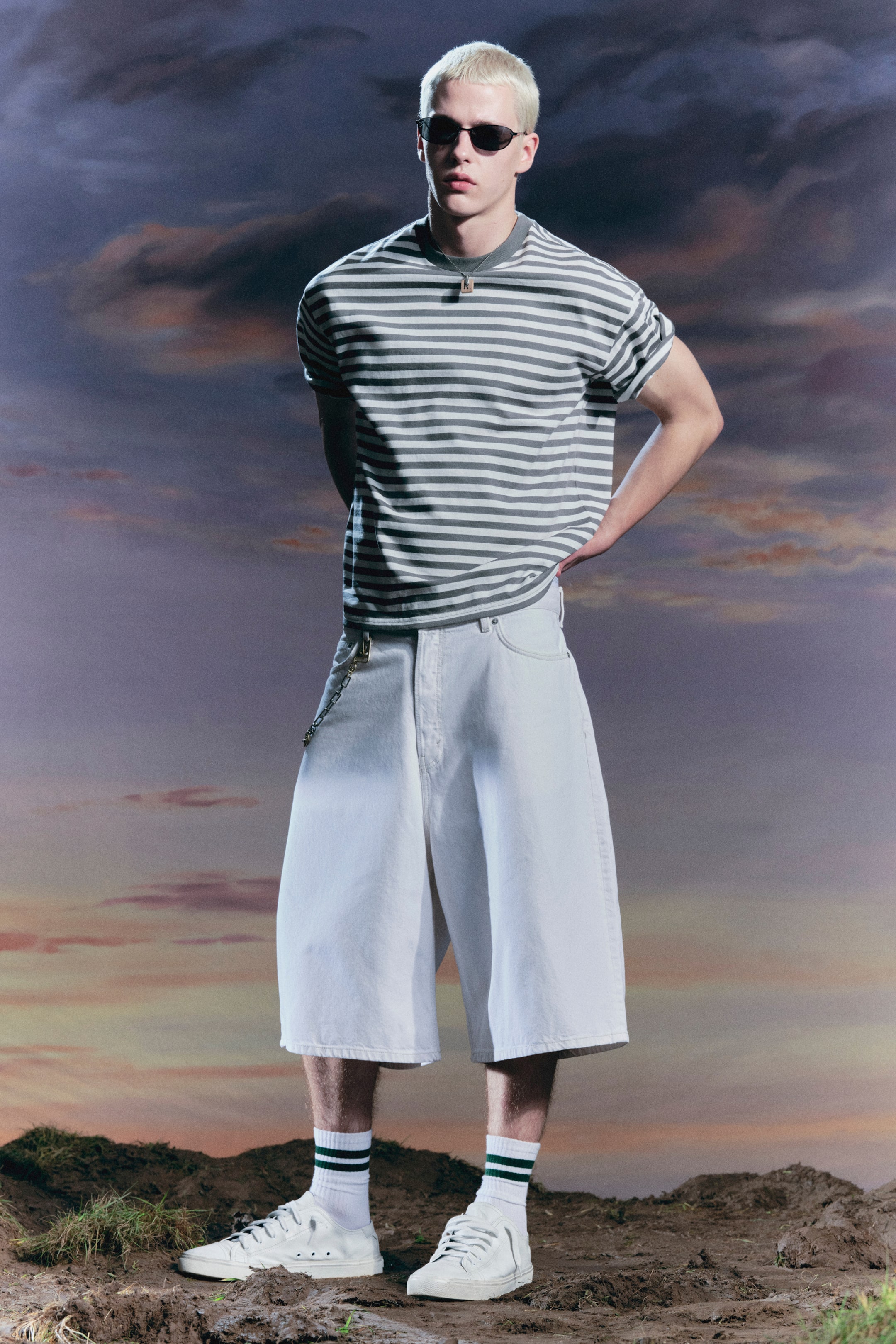 Zobraziť väčší obrázok: Sivobiele horizontálne pruhované tričko s vyhrnutými krátkymi rukávmi je nosené s svetlými, širokými šortkami, ktoré siahajú tesne pod kolená a sú ozdobené striebornou retiazkou. Biele nízke tenisky dopĺňajú vzhľad, spárované s bielymi ponožkami s dvoma zelenými pruhmi, zatiaľ čo tmavé oválne slnečné okuliare a kovový prívesok na náhrdelníku dotvárajú outfit.