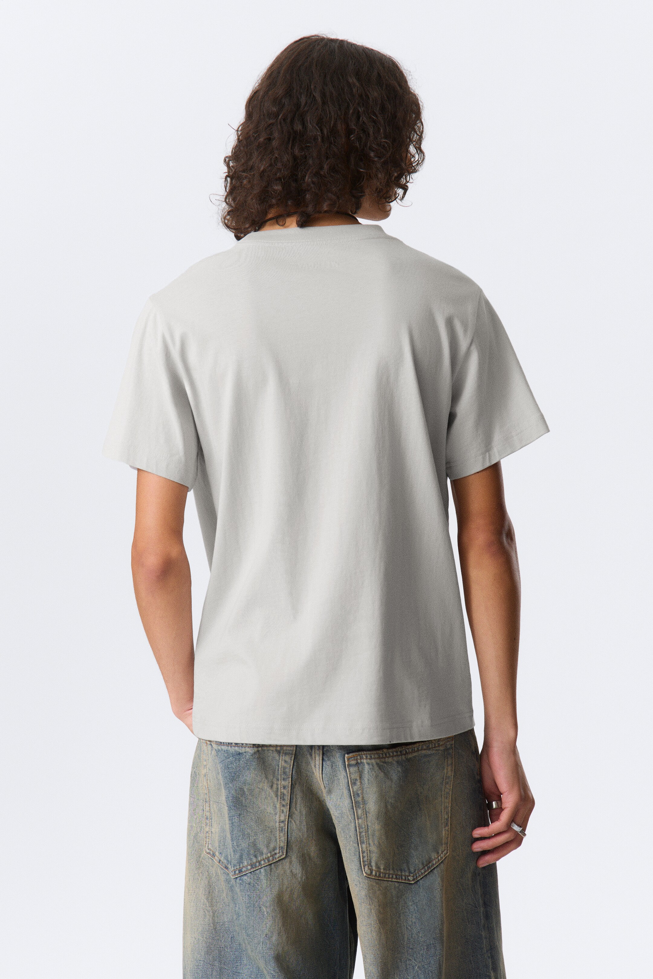 Visualizza immagine più grande: Una t-shirt grigio chiaro è presentata dalla vista posteriore, mostrando una vestibilità rilassata, maniche corte e un collo a giro arrotondato.