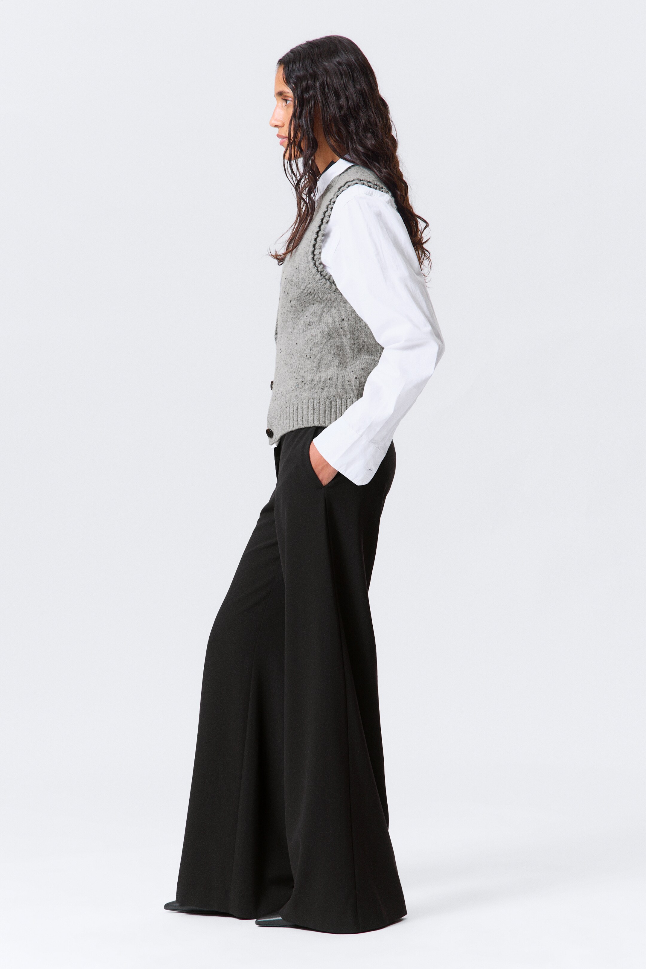 Visualizza immagine più grande: Un gilet lavorato a maglia grigio con macchie è stratificato su una camicia bianca a maniche lunghe, con entrambi gli articoli indossati con pantaloni neri a gamba larga, tutti presentati da un profilo laterale. Il gilet presenta bottoni scuri lungo la parte anteriore e un bordo smerlato lungo lo scollo a V e i fori per le braccia, mentre i pantaloni sono progettati con tasche laterali e una silhouette fluente.