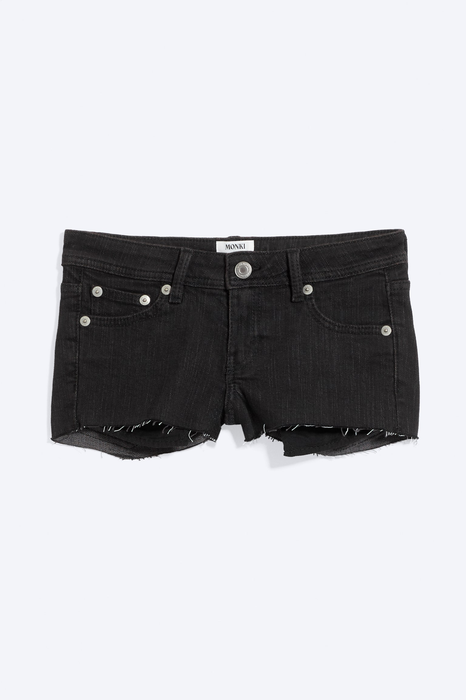 Pantaloncini corti in denim nero, a vita bassa e vestibilità slim, sono mostrati frontalmente, con chiusura a bottone e zip, due tasche anteriori con rivetti in metallo, una tasca portamonete e un orlo grezzo e sfrangiato. Un'etichetta 'MONKI' è visibile al centro della cintura superiore.