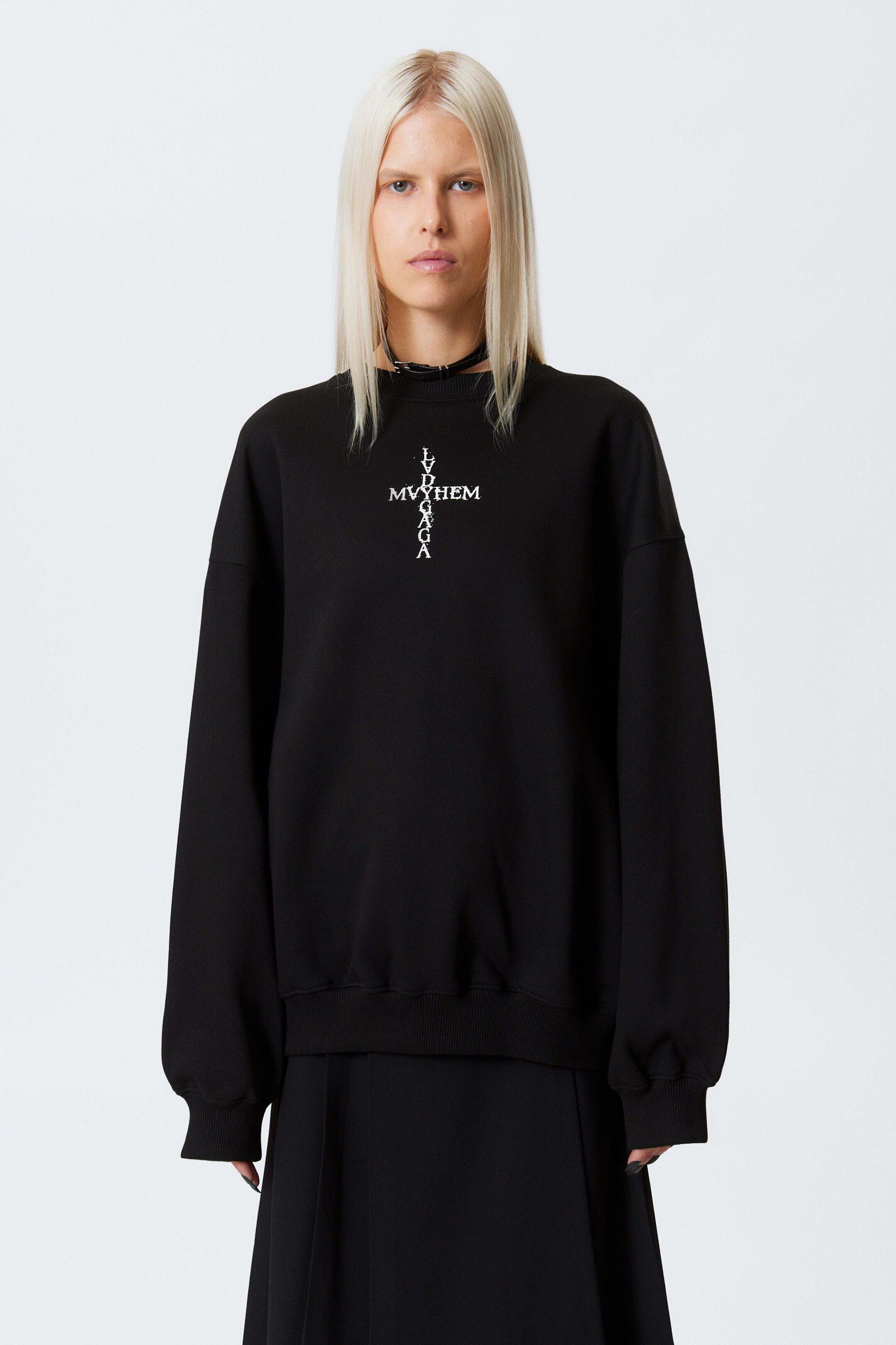 Visualizza immagine più grande: Una felpa nera oversize viene presentata, caratterizzata da un design grafico a croce bianca sul petto con "LV MYYHEM" in orizzontale e "LVDGAGA" in verticale, entrambi con una texture consumata. È progettata con un girocollo, polsini a coste e un orlo a coste, ed è indossata sopra una gonna nera plissettata con un choker nero visibile al collo.
