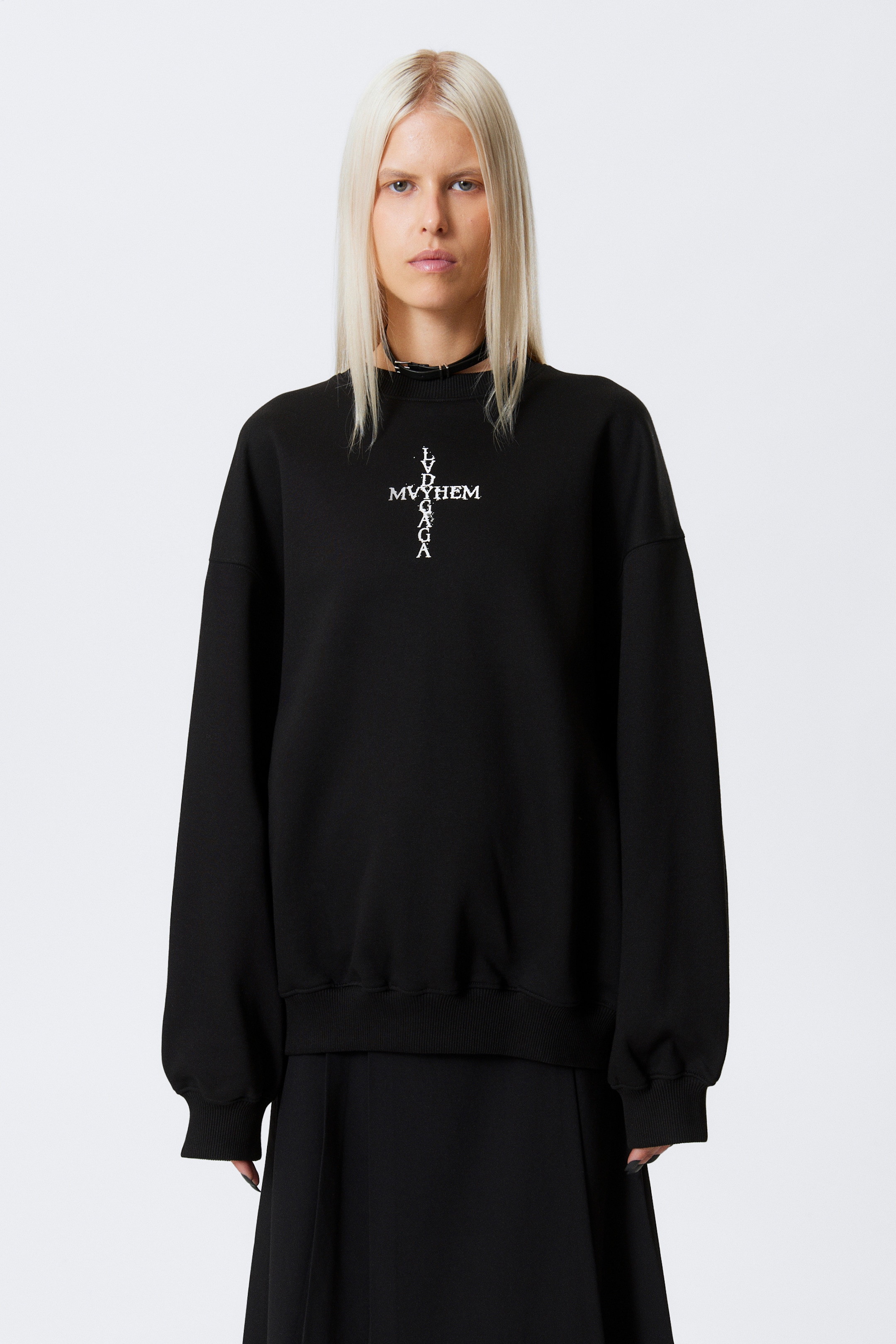 Agrandir l'image: Un sweat-shirt noir oversize est présenté, avec un motif croix graphique blanc sur la poitrine portant les inscriptions "LV MYYHEM" horizontalement et "LVDGAGA" verticalement, tous deux avec une texture usée. Il est conçu avec un col rond, des poignets côtelés et un ourlet côtelé, et est porté sur une jupe plissée noire avec un collier ras du cou noir visible au niveau du col.
