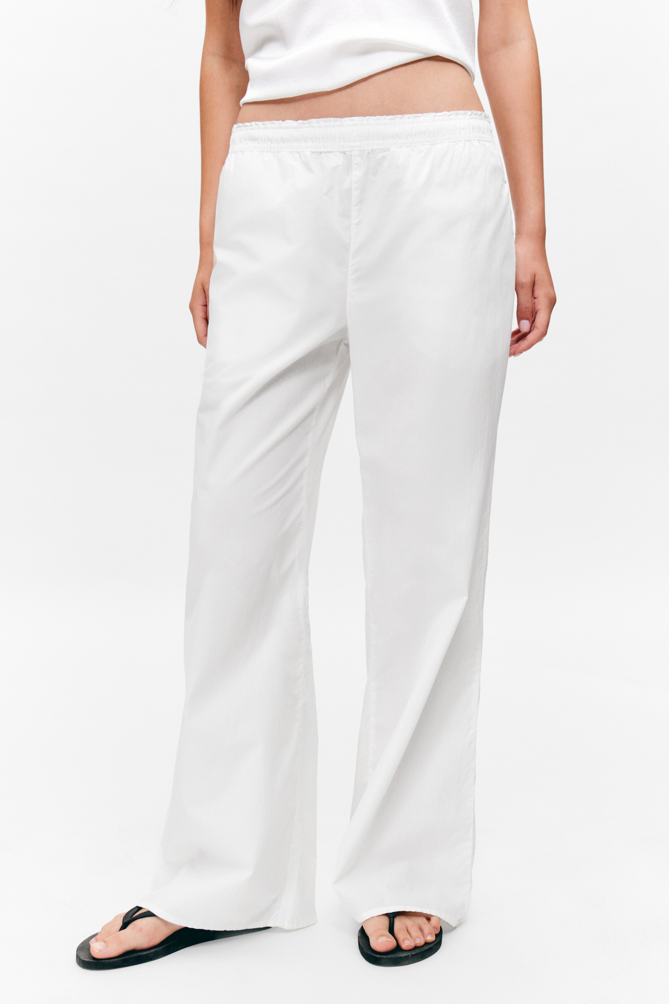 Größeres Bild anzeigen: Weiße Hosen mit weitem Bein werden gezeigt, die über einen elastischen, gerafften Bund verfügen. Die Hosen zeigen eine entspannte Passform, wobei der Stoff locker bis zum Boden fließt und teilweise die Füße des Trägers bedeckt.