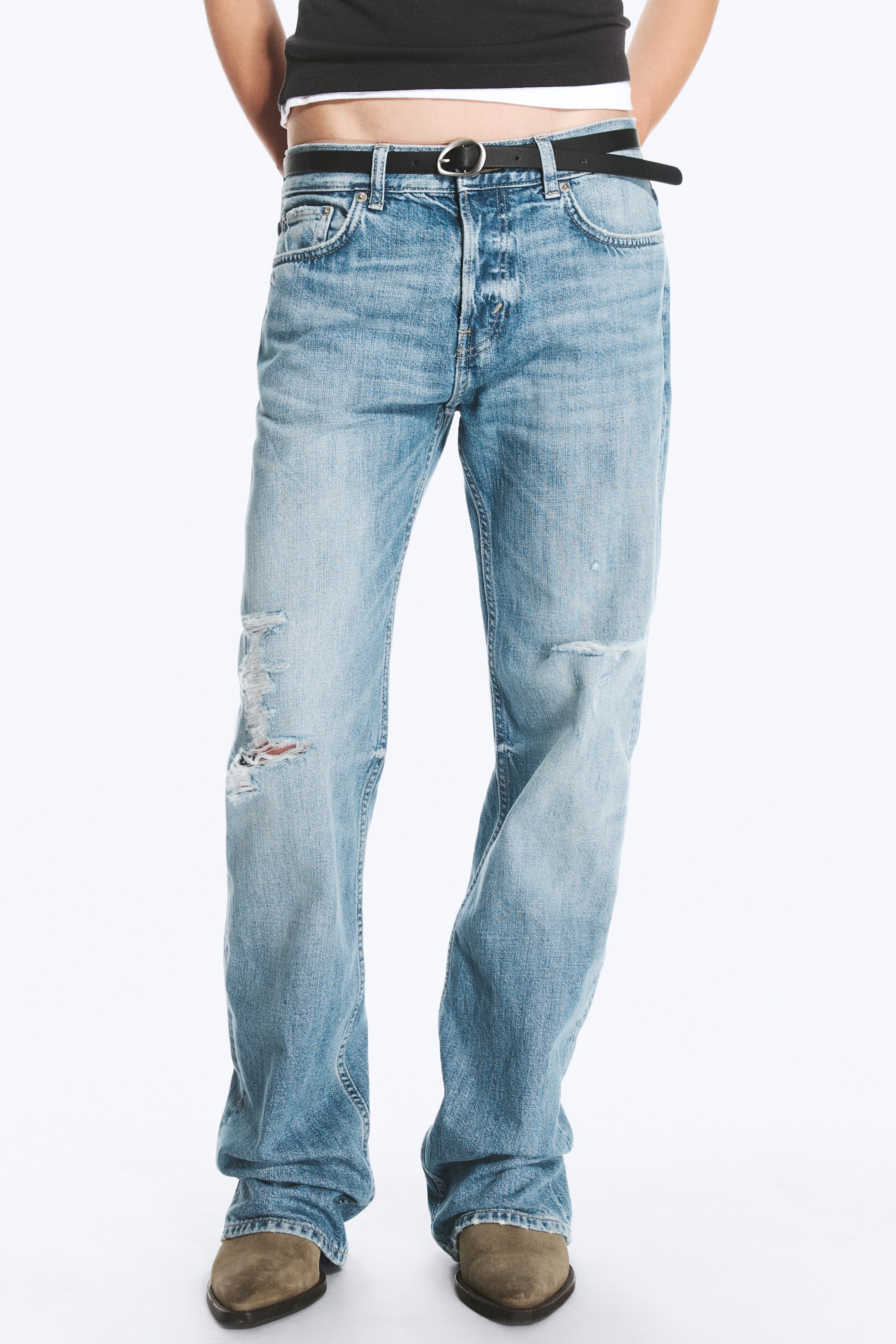 Agrandir l'image: Le mannequin porte un jean droit regular taille basse Sonic en délavage bleu clair. Ce jean présente des détails vieillis aux genoux et est associé à une ceinture en cuir noir, un top noir court et des bottes marron.