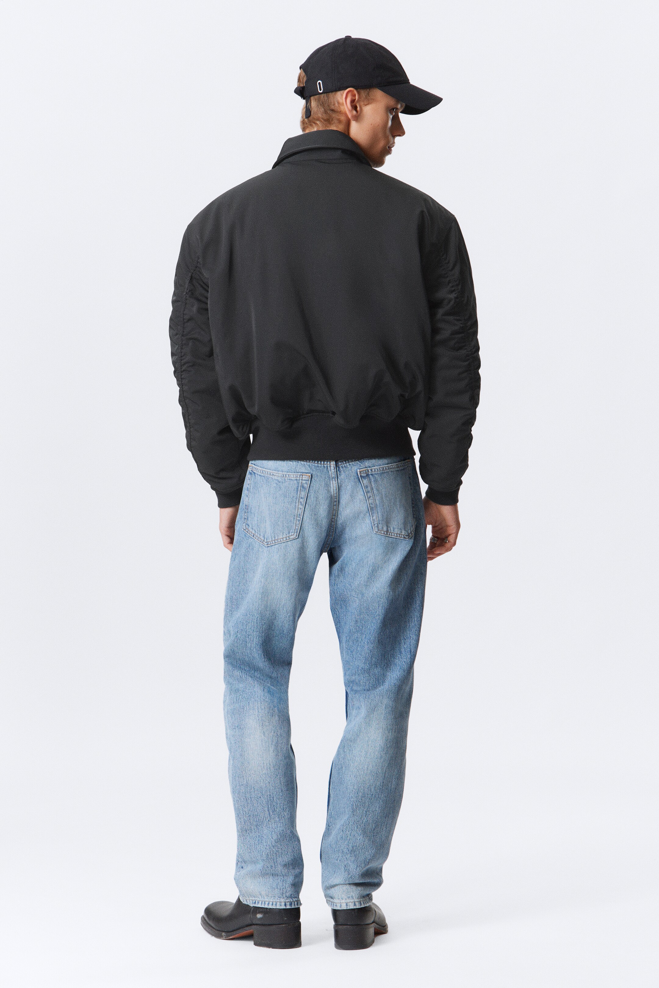 Visualizza immagine più grande: Una giacca bomber nera presenta un colletto a punta, maniche ampie e arricciate e un orlo a coste, ed è indossata con jeans in denim chiaro. I jeans presentano una vestibilità rilassata e due tasche posteriori applicate, con l'intero ensemble visto da dietro.