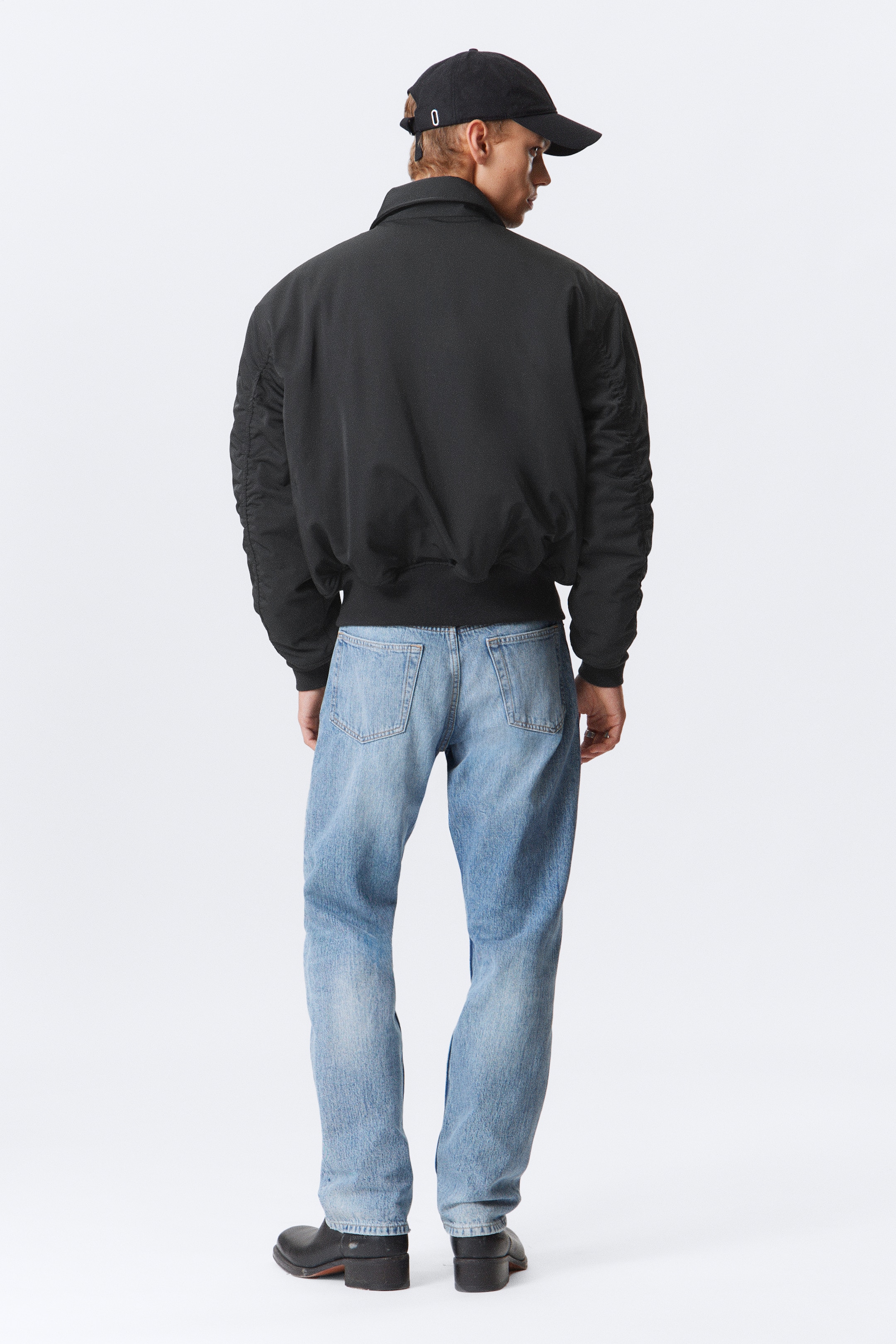 Visa större bild: En svart bomberjacka har en spetsig krage, voluminösa rynkade ärmar och en ribbad fåll och bärs tillsammans med ljusa denimjeans. Jeansen har en avslappnad passform och två bakre påfickor, och hela outfiten betraktas bakifrån.
