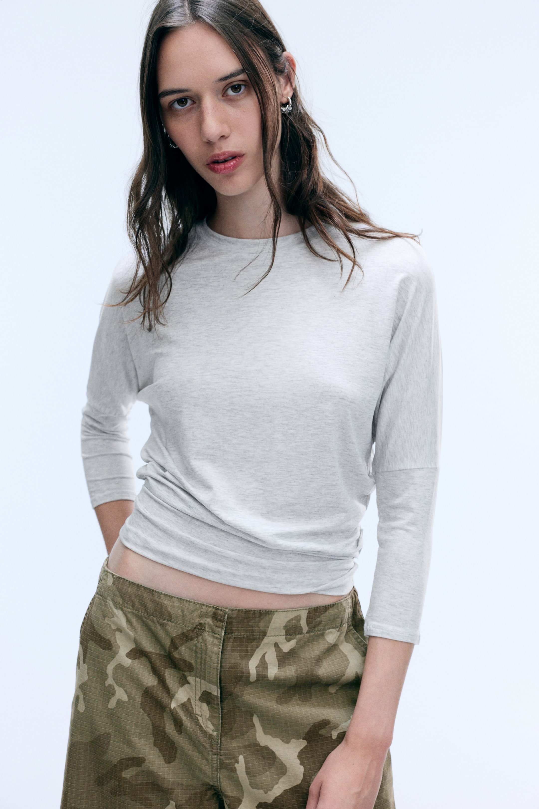Ver imagen más grande: La modelo lleva un top gris claro de manga tres cuartos con cuello redondo, hombros caídos y fruncido drapeado en la cintura. El top se combina con un pantalón de estampado de camuflaje.
