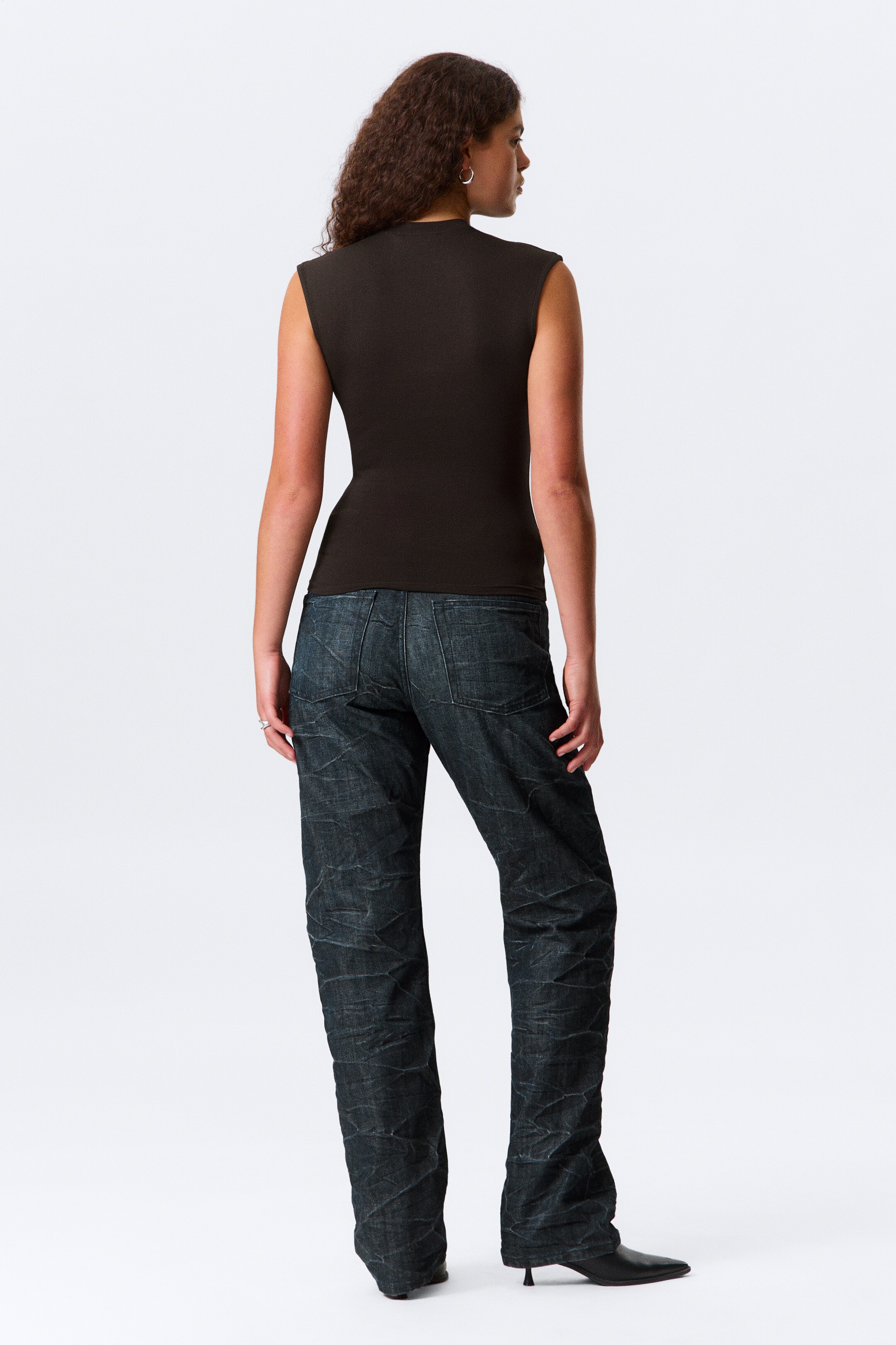 Größeres Bild ansehen: Ein dunkelbraunes ärmelloses Top wird von hinten gezeigt, leicht nach links, eng anliegend am Körper. Es wird mit dunklen, gerade geschnittenen Jeans kombiniert, die eine sichtbare geknitterte Textur aufweisen und über schwarzen, spitzen Kitten-Heel-Stiefeln enden.