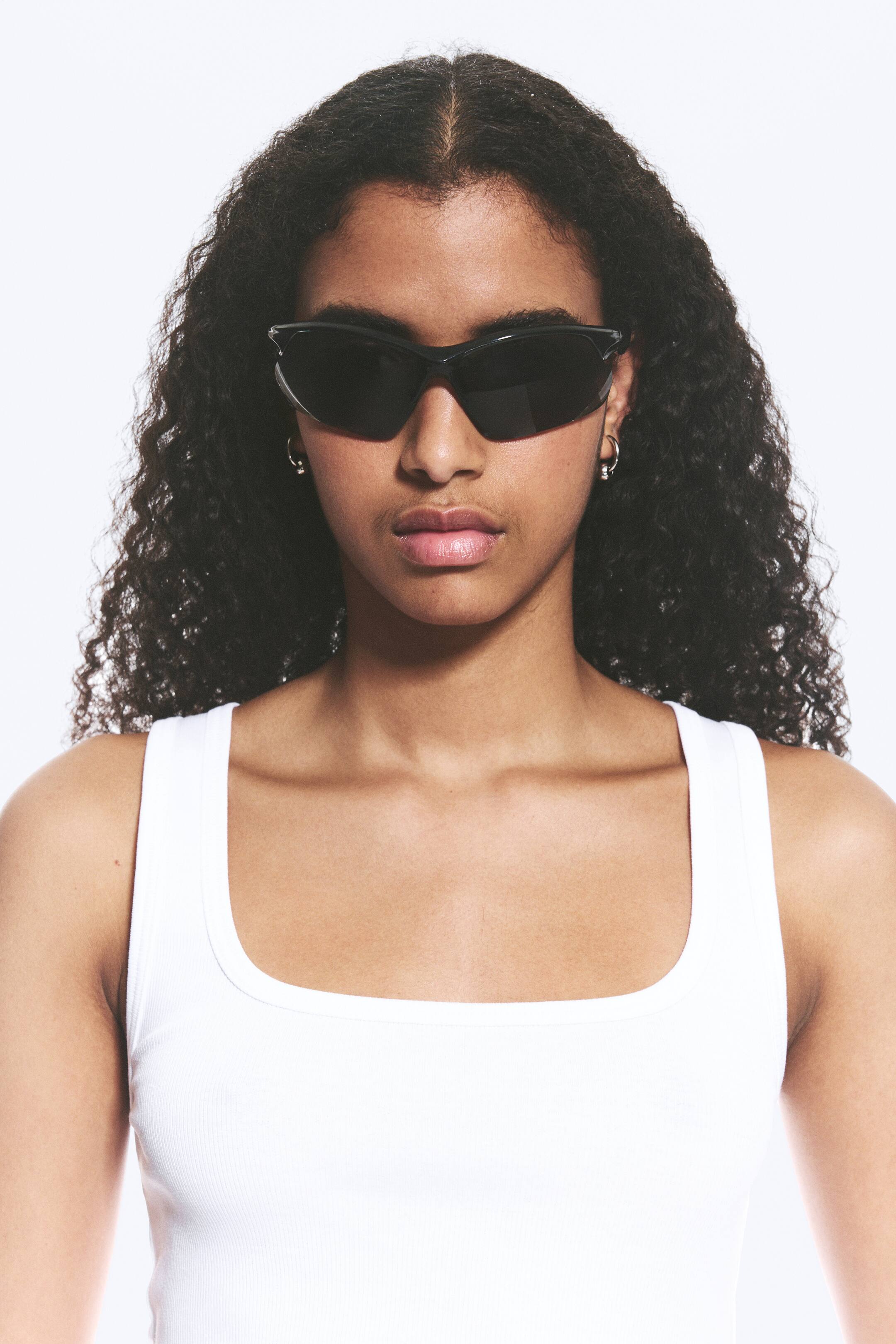 View larger image: The model is wearing black, futuristic sunglasses featuring tinted lenses and a streamlined frame with slender temples.