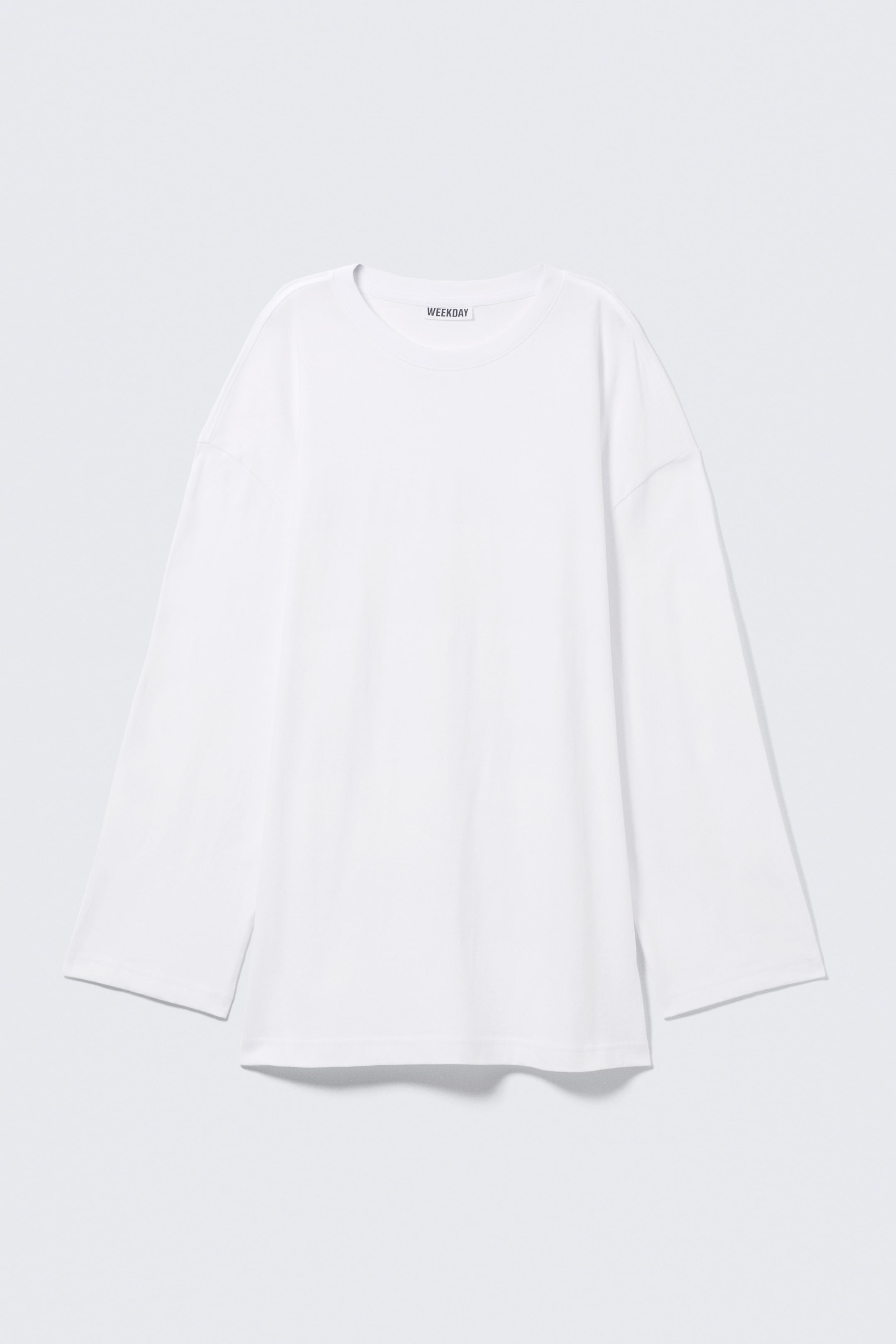 Se større billede: En hvid langærmet t-shirt vises som FRONT, med en oversized, afslappet pasform. Den har en rund halsudskæring, brede dropped-shoulder ærmer og et synligt "WEEKDAY" mærke i den indvendige halsudskæring.