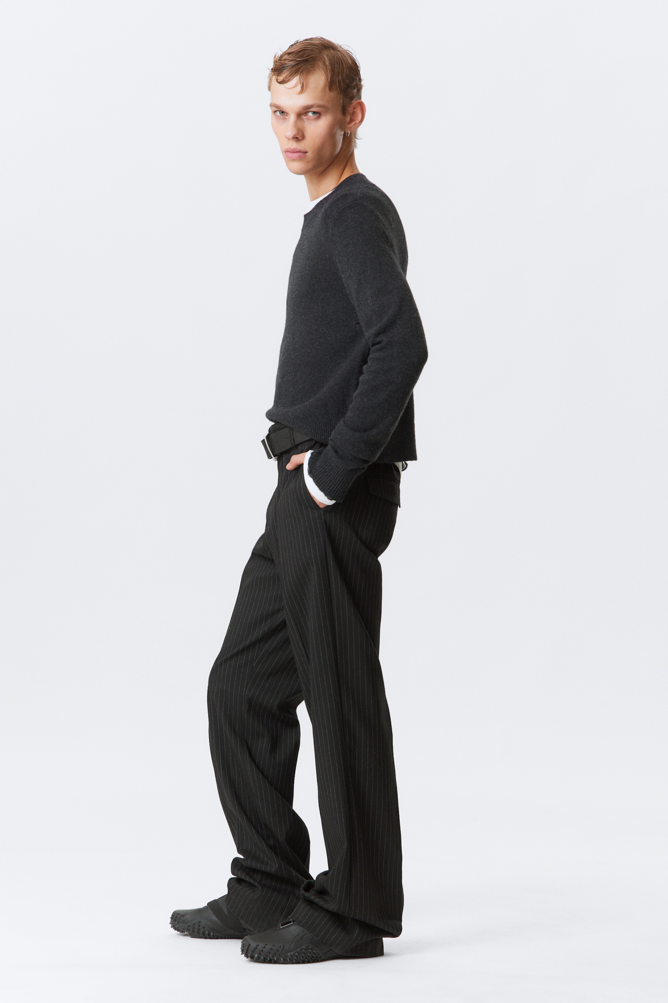 Agrandir l'image: Un pull à col rond gris foncé est présenté d'un léger angle de côté, montrant sa coupe décontractée et présentant des poignets et un ourlet côtelés, avec un poignet blanc visible en dessous. Un pantalon à fines rayures foncées avec une coupe large est porté, sécurisé par une ceinture noire, le tissu s'accumulant sur des chaussures noires sans lacets dotées d'une semelle texturée.