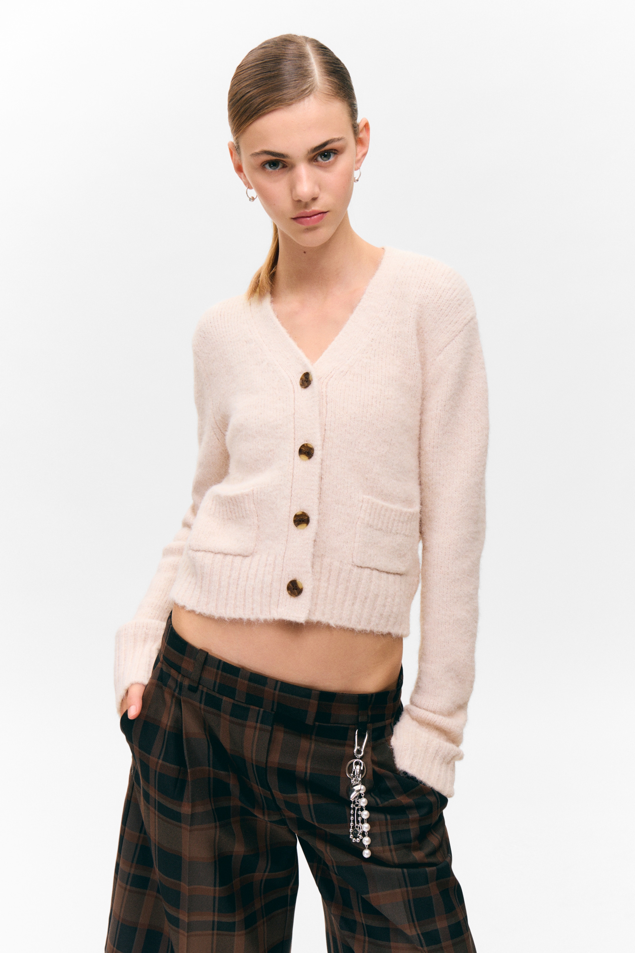 Grösseres Bild ansehen: Eine hellfarbene, weiche Strickjacke mit V-Ausschnitt, Knopfleiste mit vier sichtbaren Schildpattknöpfen und zwei aufgesetzten Taschen vorne. Sie hat lange Ärmel mit gerippten Bündchen und einen gerippten, verkürzten Saum, alles in einer entspannten Passform aus der Vorderansicht.