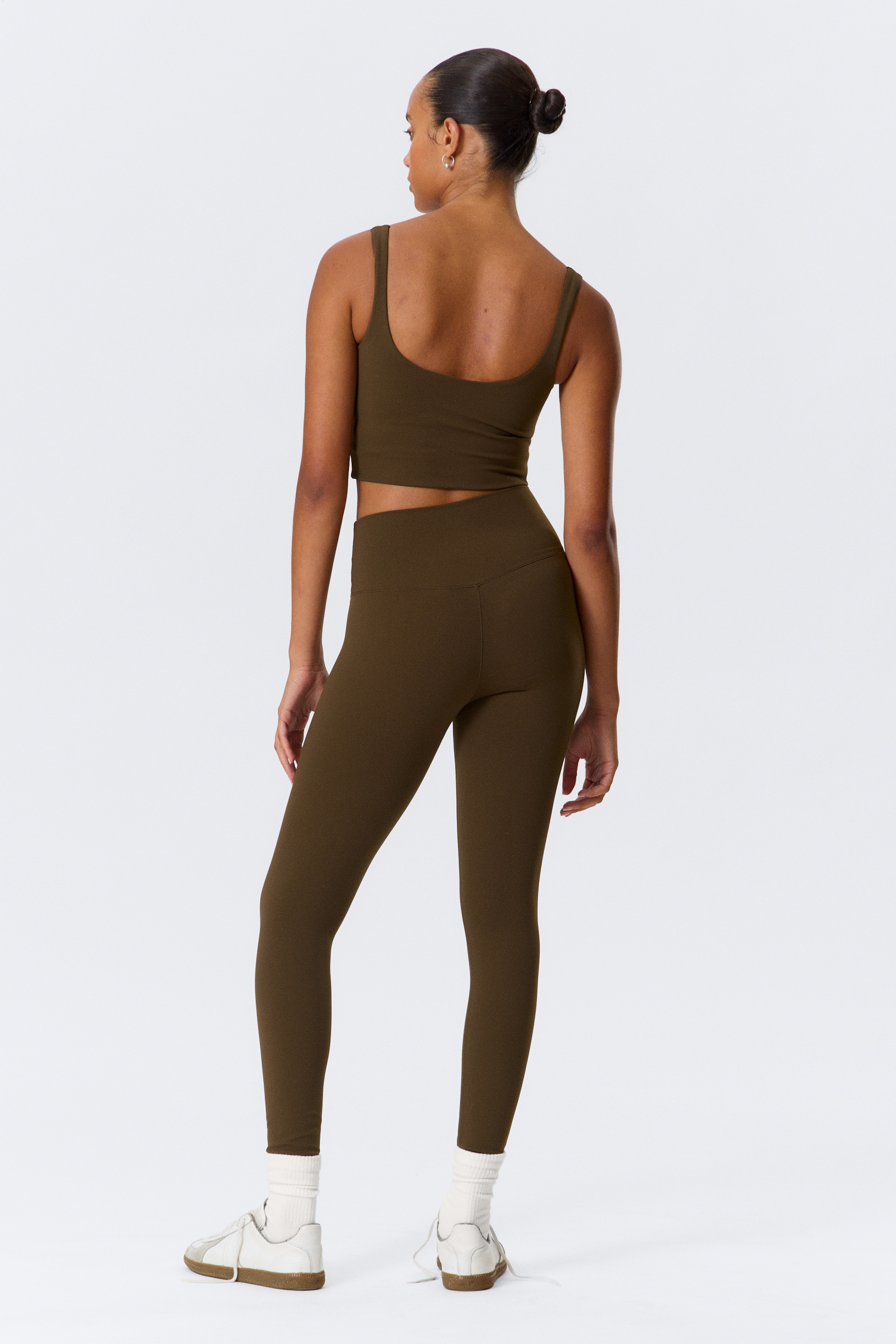 Größeres Bild anzeigen: Ein sportliches Set in dunkelolivgrün wird aus einem hinteren Winkel gezeigt. Das Set besteht aus einem kurzen Tanktop mit breiten Schulterträgern und einem tiefen Rückenausschnitt, kombiniert mit hoch taillierten Leggings, die eine V-förmige Naht am hinteren Bund aufweisen.