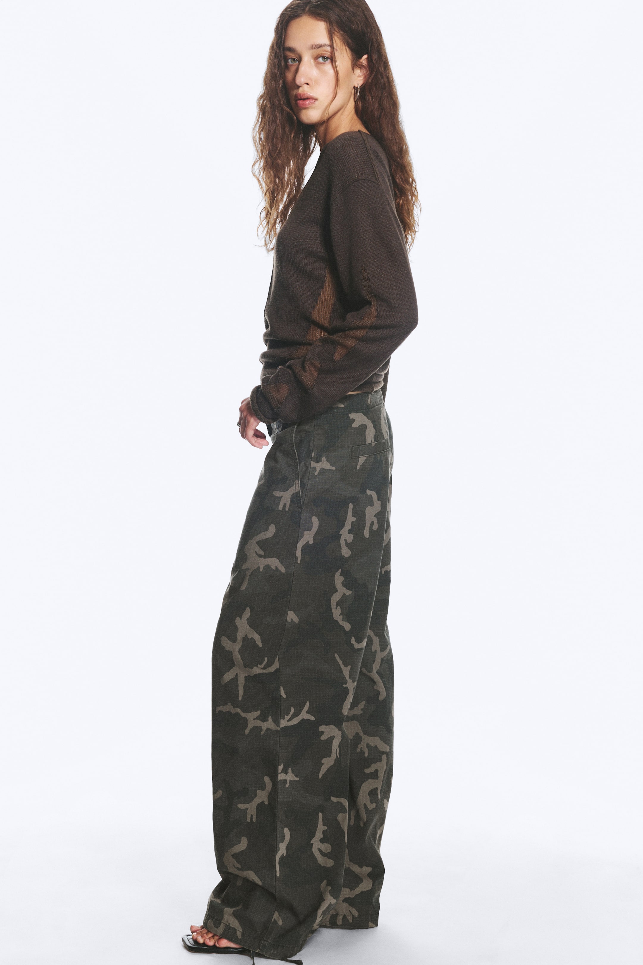 View larger image: A pair of low-rise trousers, featuring a camouflage pattern and a loose, baggy leg silhouette, is being worn by the model. The trousers have cinched hems.