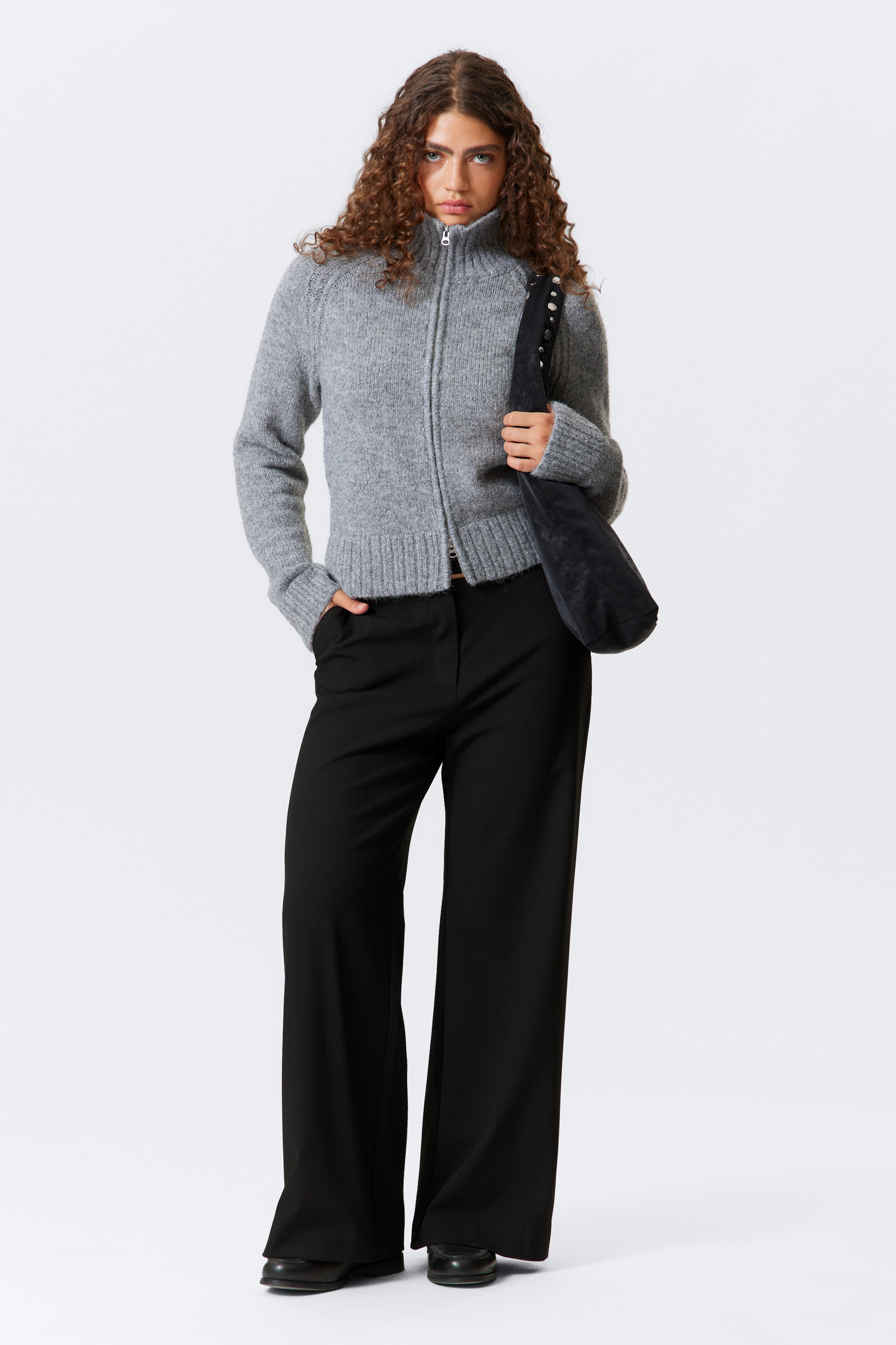 Grösseres Bild ansehen: Ein graues gestricktes Zip-up-Oberteil zeigt einen Stehkragen, Raglanärmel und gerippte Bündchen und einen gerippten Saum. Es wird mit schwarzen Hosen mit weitem Bein kombiniert.