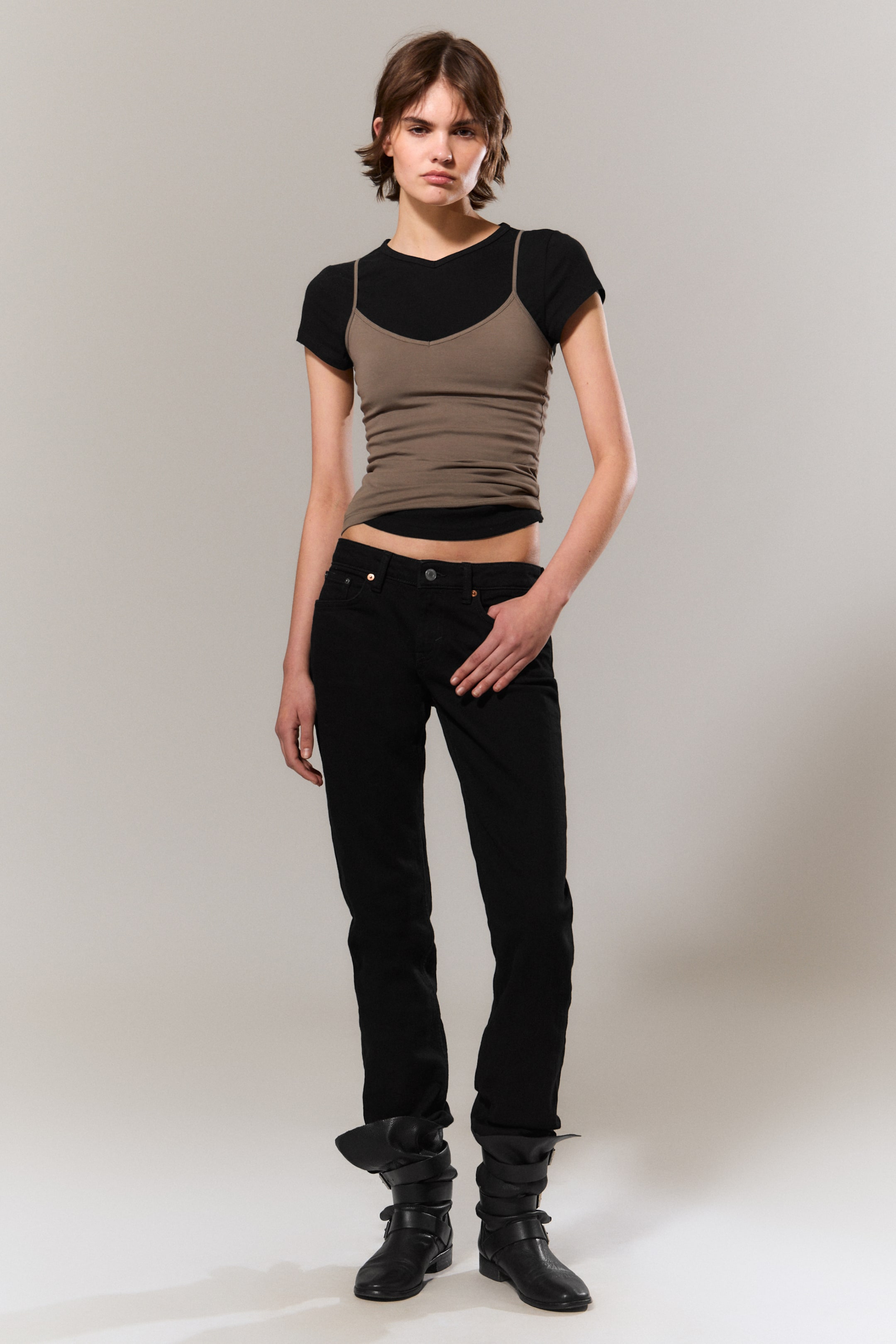 Zobraziť väčší obrázok: Outfit predstavuje čierne tričko s V-výstrihom, ktoré je vrstvené pod priliehavým hnedým tielkom s V-výstrihom a špagetovými ramienkami. Súprava je doplnená čiernymi slim-fit džínsami, ktoré sú vyhrnuté nad čierne členkové topánky s prackovými remienkami.