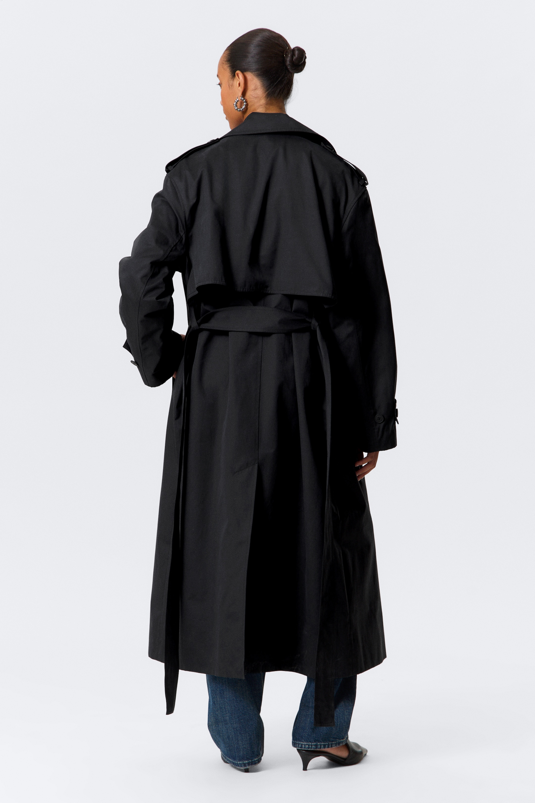 Visualizza immagine più grande: Un trench coat nero viene mostrato da dietro, estendendosi fino al polpaccio. Presenta spalline sulle spalle, un'ampia pattina antipioggia sulla parte superiore della schiena e maniche lunghe con cinturini regolabili ai polsi. Una cintura in tessuto coordinato stringe la vita, con le sue estremità lunghe che pendono sopra una spacco centrale sul retro.