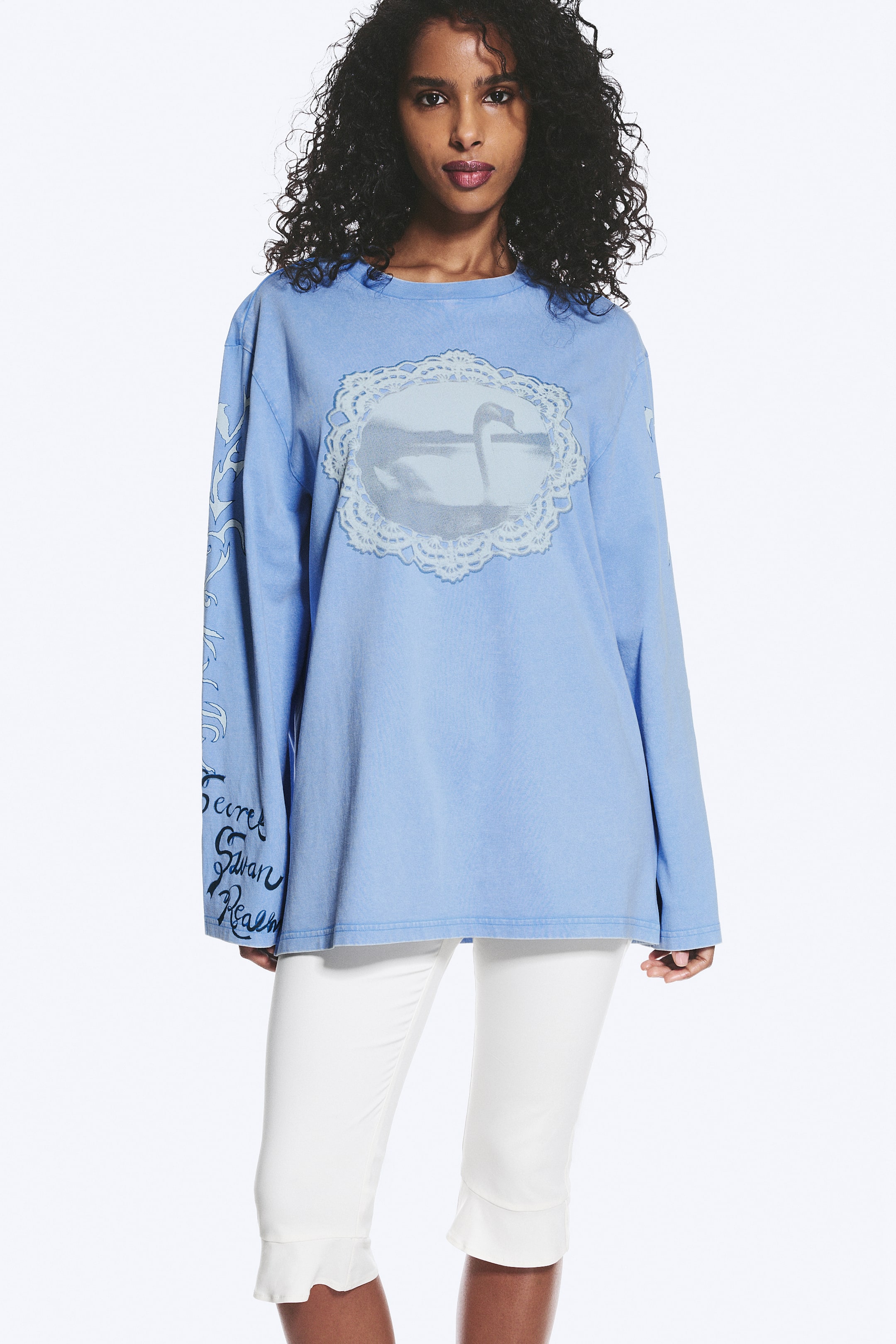 Visa större bild: En ljusblå långärmad t-shirt med rund hals har en oversized passform och ett tvättat utseende. Framsidan visar en central oval grafik av en svan på vatten, inramad av en vit spetsliknande kant, medan vänster ärm pryds av svart skripttext och dekorativa utsmyckningar.