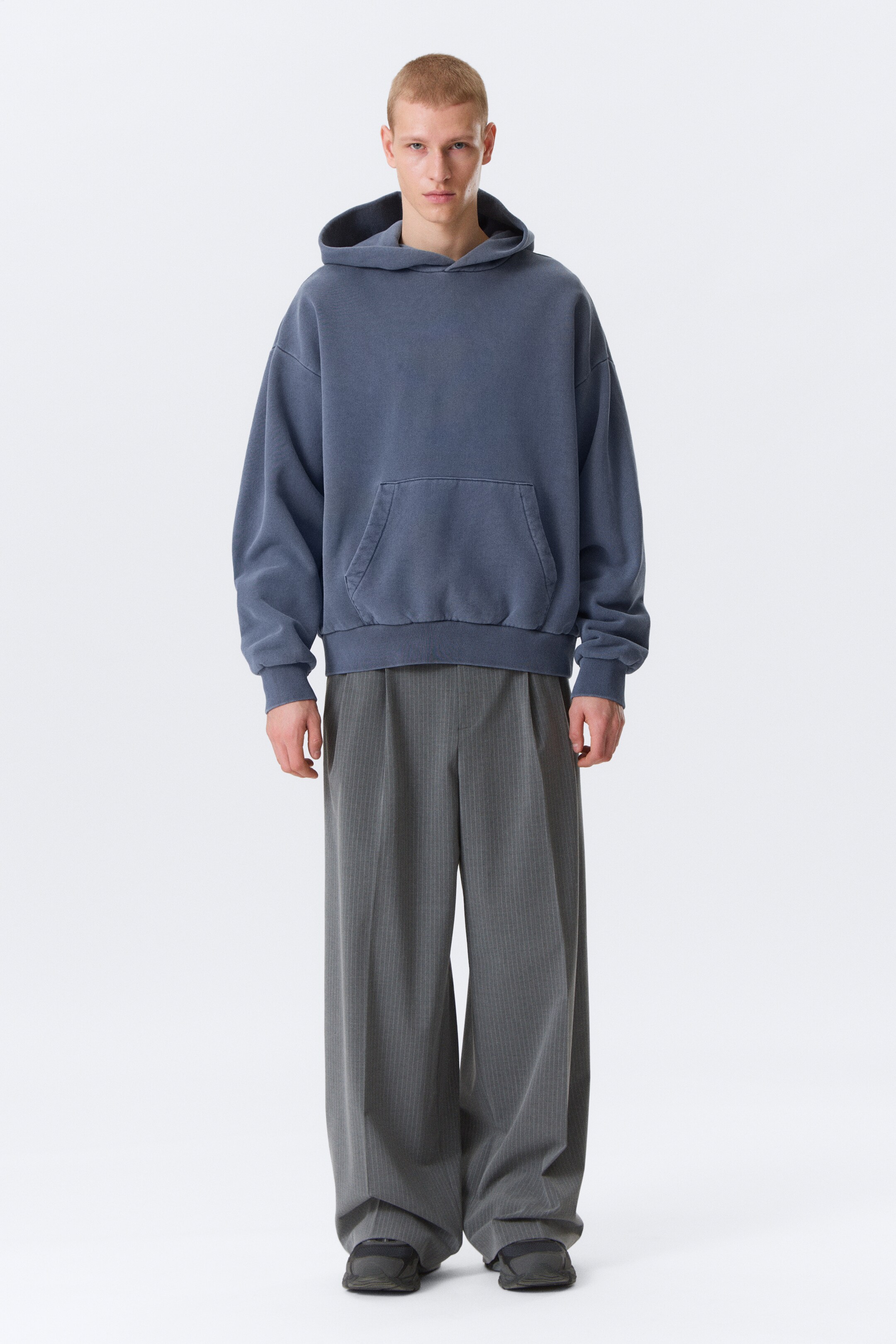 Ingrandisci l'immagine: Una felpa oversize color blu-grigio sbiadito è presentata, caratterizzata da un cappuccio con coulisse, una tasca frontale a marsupio e polsini e orlo a coste. È indossata con pantaloni a gamba larga color grigio scuro, che mostrano sottili righe verticali e pieghe frontali.