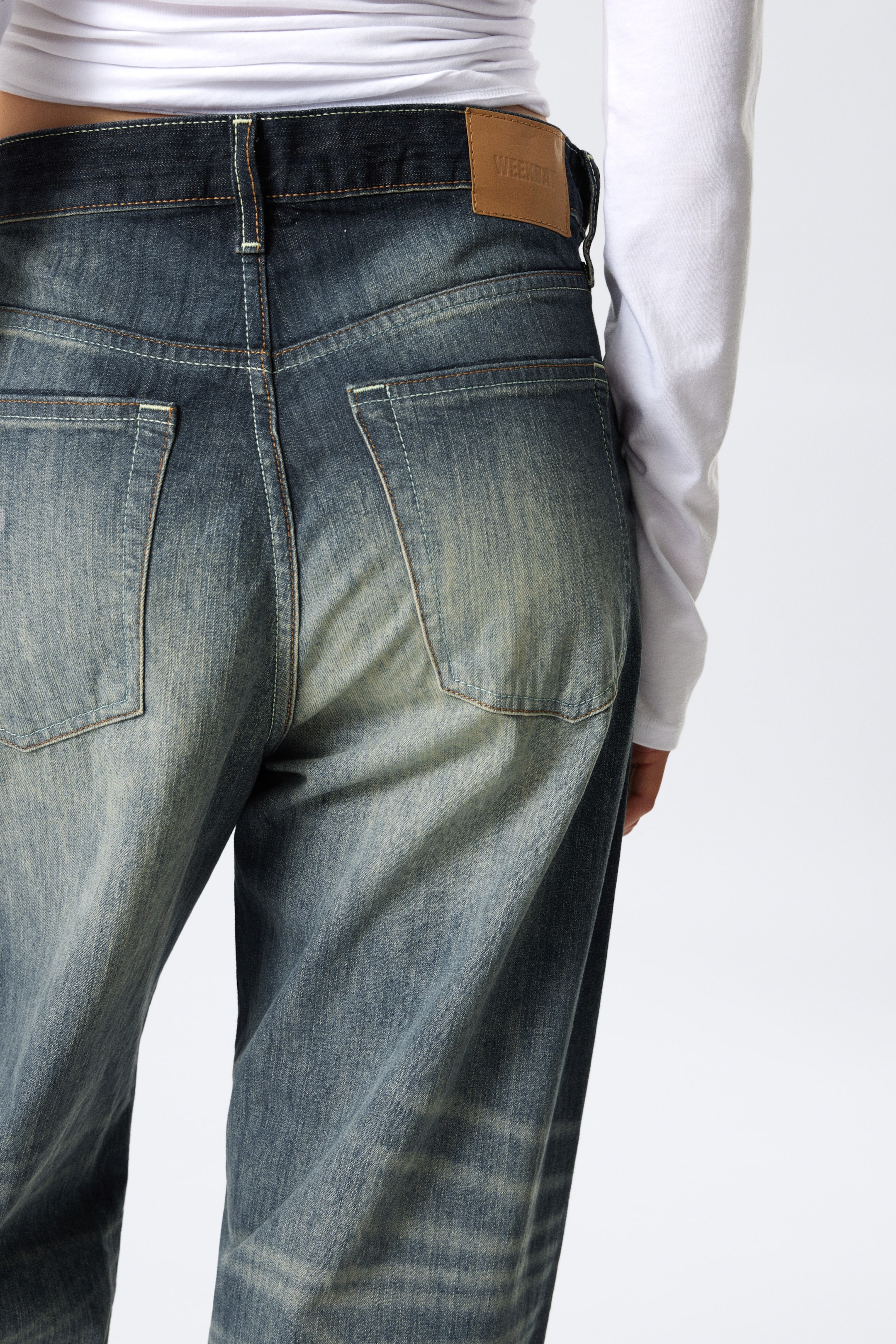 View larger image: Dark blue denim jeans are being displayed from the back, featuring a faded wash with lighter areas on the seat and thighs. Contrast stitching is accentuating the seams and rear patch pockets, and a light brown brand patch is visible on the right side of the waistband. A white long-sleeved top is tucked into the jeans, illustrating the high-waisted fit.