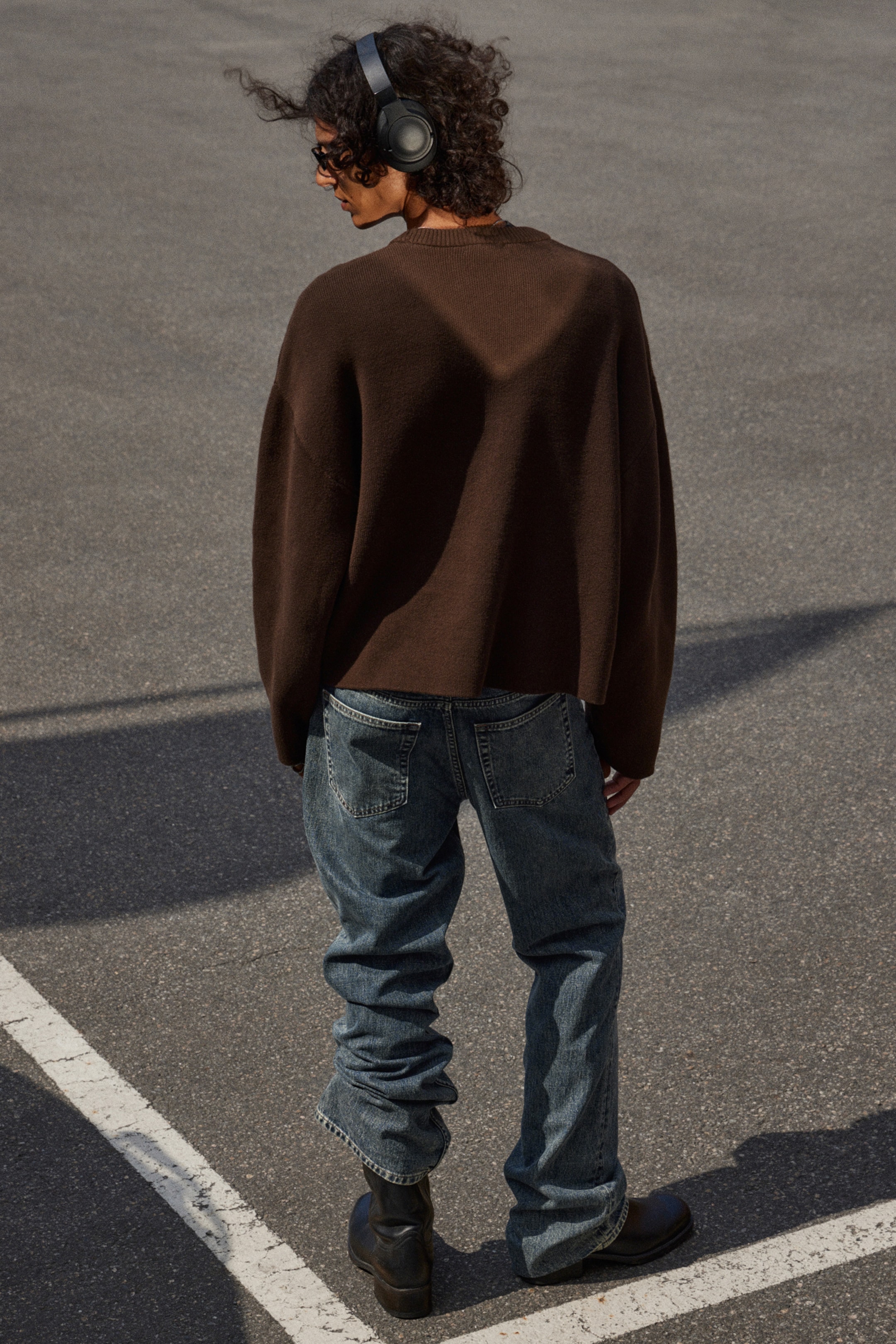 Größeres Bild anzeigen: Ein übergroßer dunkelbrauner Strickpullover wird getragen, mit einer entspannten, langärmeligen Passform. Er wird mit locker sitzenden, hell gewaschenen blauen Jeans kombiniert, die sichtbar an den Knöcheln über schwarzen Stiefeln gestapelt sind, alles von hinten betrachtet.