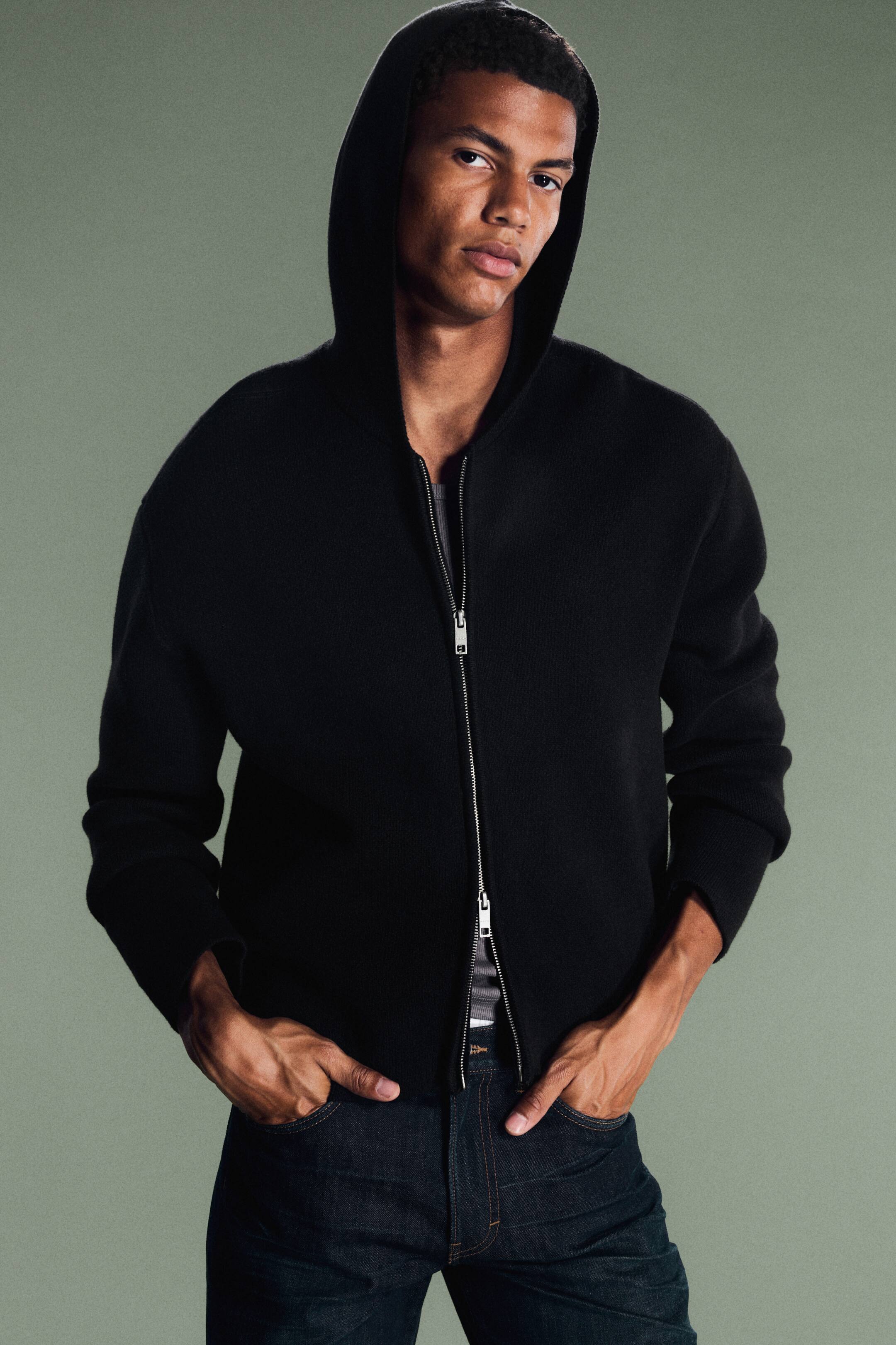 Visualizza immagine più grande: Una felpa con cappuccio nera in maglia, con zip integrale, indossata dalla modella con il cappuccio tirato su sulla testa. La felpa presenta una cerniera bidirezionale argentata, parzialmente aperta per mettere in evidenza un sotto-strato grigio, ed è abbinata a jeans dal lavaggio scuro.
