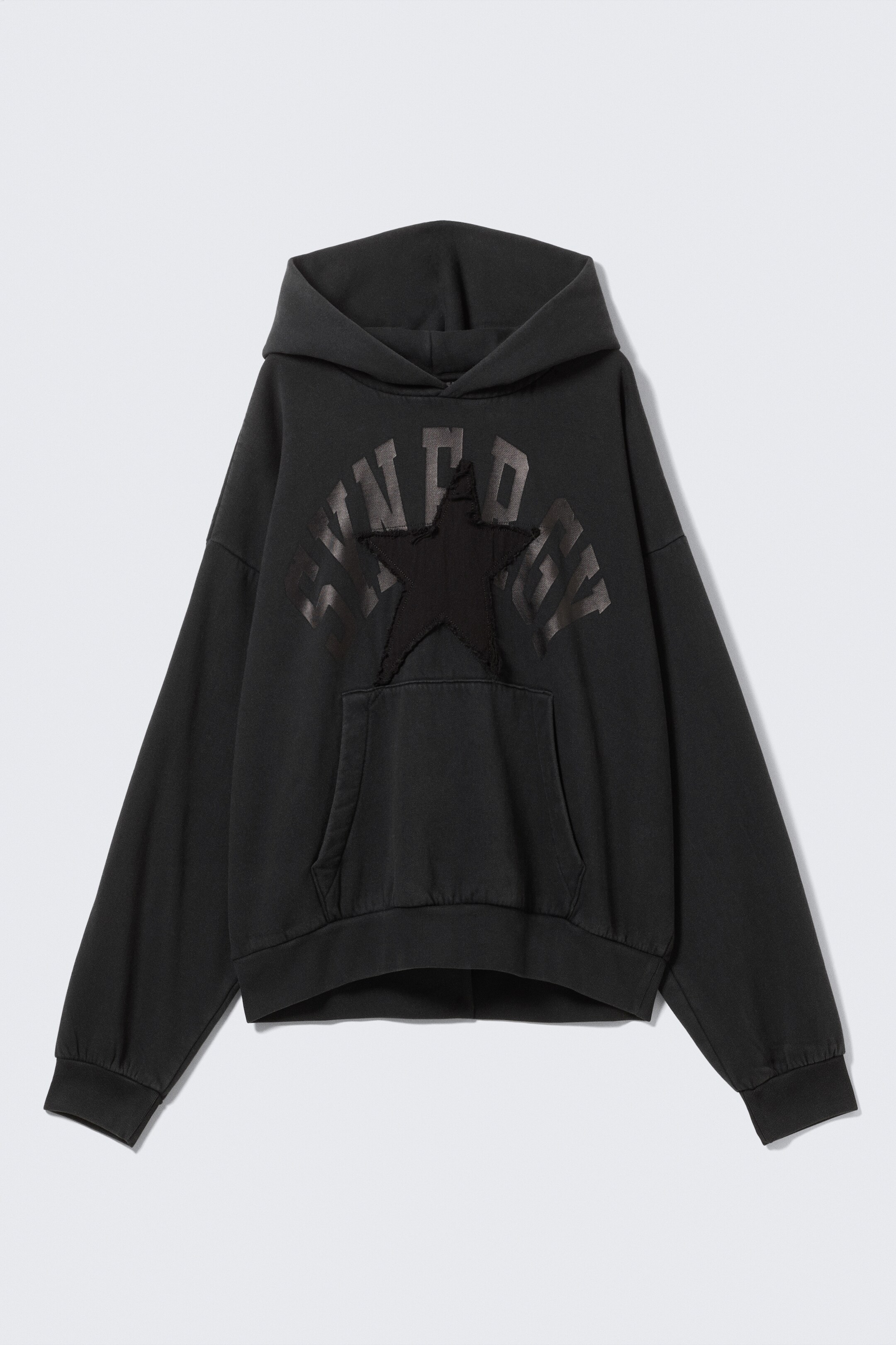 View larger image: A loose-fit black graphic hoodie, crafted from soft cotton fleece, is being presented from the FRONT, featuring dropped shoulders, a roomy hood, and gathered hems. A large, subtle dark grey graphic, comprising distressed text and a central star patch, is visible on the chest above a kangaroo pocket, with ribbed trims finishing the cuffs and hem.