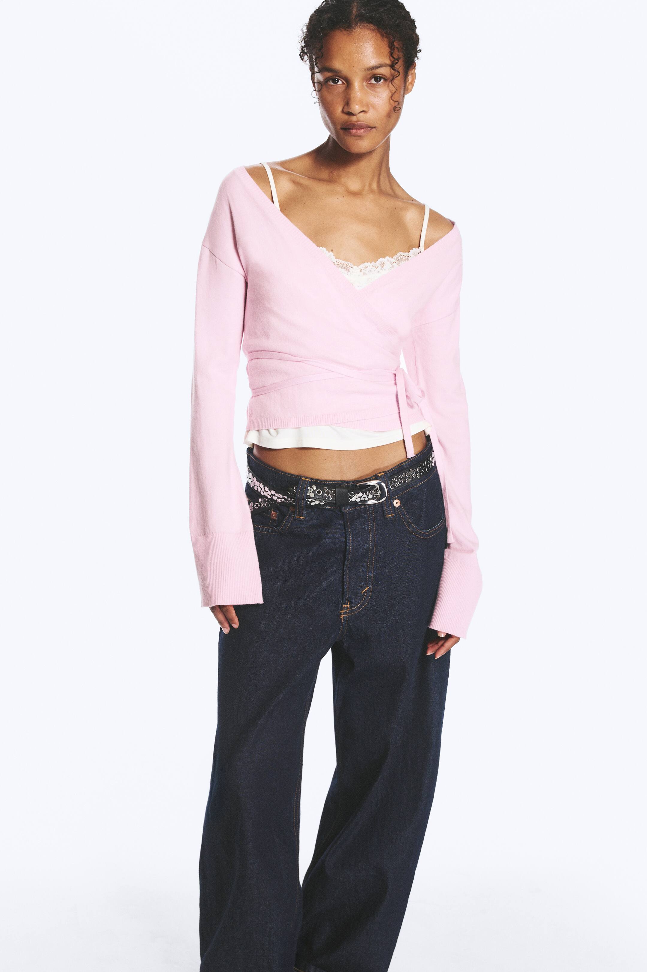 Agrandir l'image: Un cardigan en maille rose pâle à fermeture croisée est porté par le mannequin, avec un profond décolleté en V et une fermeture à nouer sur le côté. Le cardigan est superposé à un caraco blanc bordé de dentelle et associé à un jean bleu foncé.