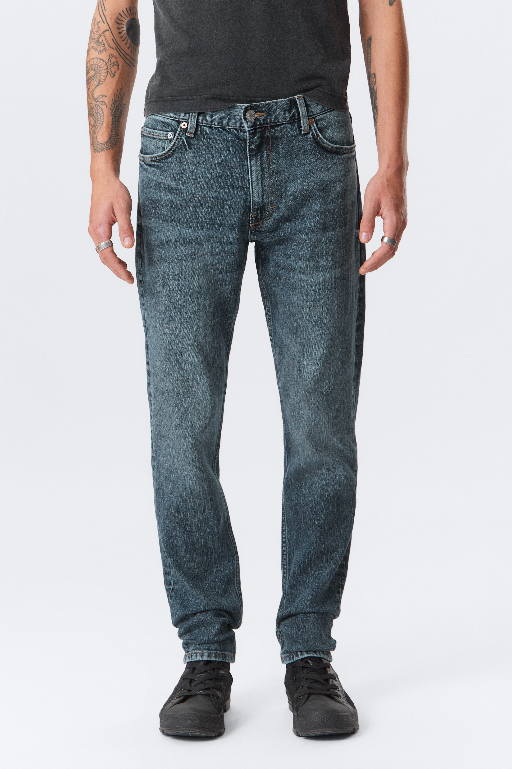 Agrandir l'image: Un jean en denim bleu-gris foncé est présenté de face, arborant un délavage dégradé avec des plis moustache sur les cuisses. Ils sont conçus avec un style classique à cinq poches, une braguette à fermeture éclair avec un bouton et des passants de ceinture, présentant une coupe droite qui repose sur des baskets noires.