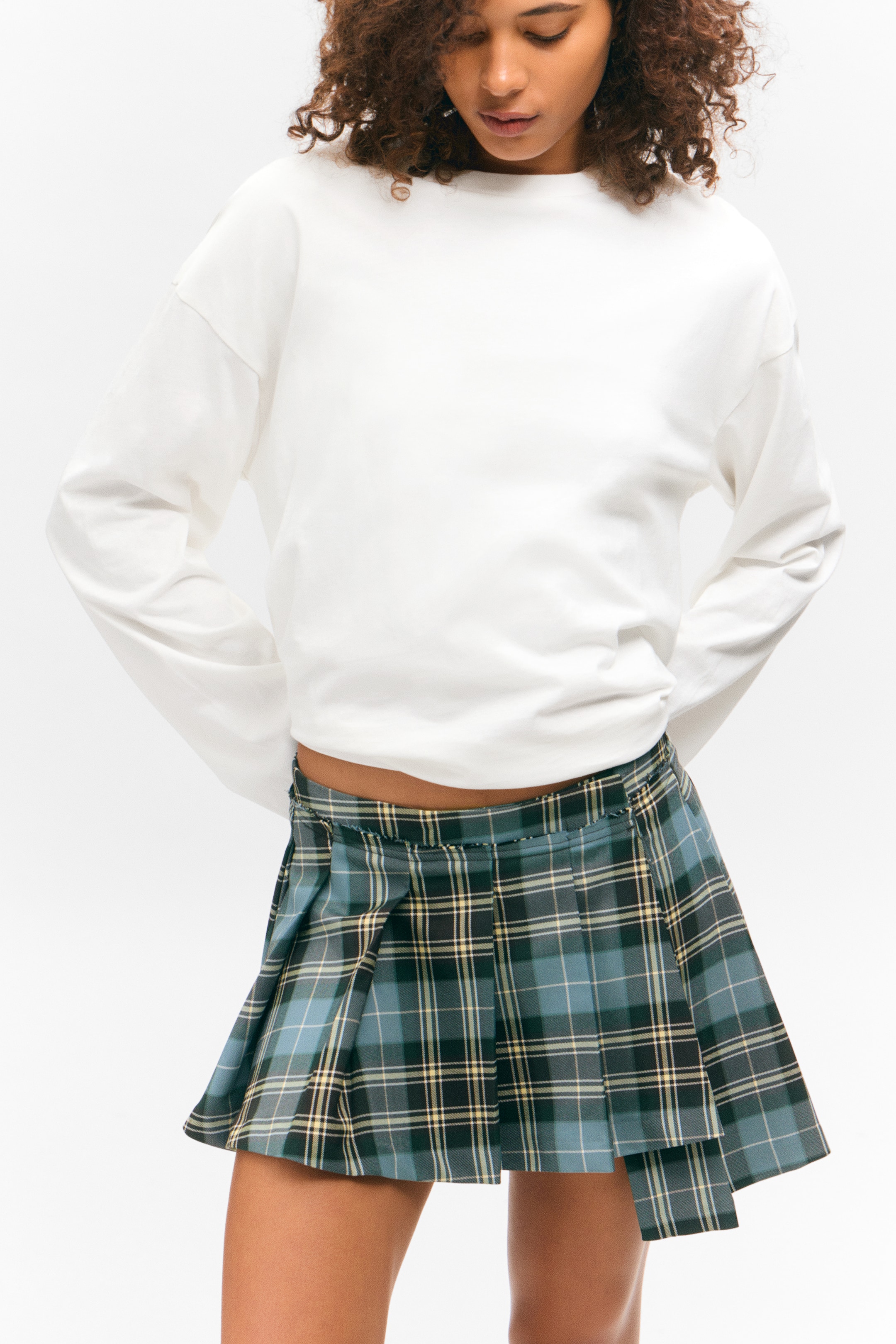 View larger image: A white long-sleeved crew-neck top is appearing with a relaxed, slightly cropped fit. Below it, a pleated mini skirt is displaying a teal, dark green, and yellow plaid pattern, with its pleats fanning out slightly as it faces forward.