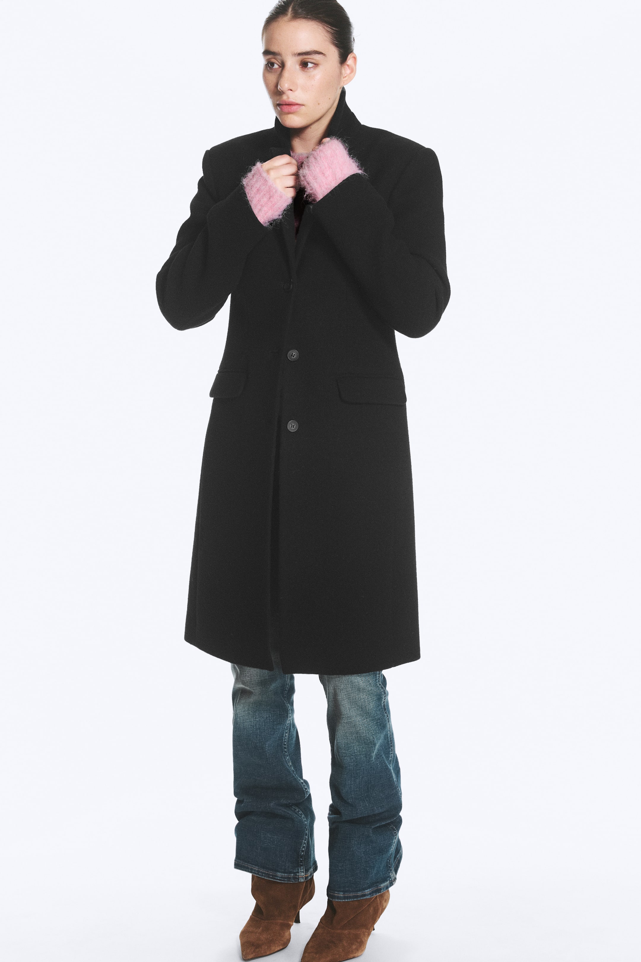 View larger image: A black, knee-length coat featuring a structured collar, front button fastening and side flap pockets is worn by the model. The model is adjusting the collar with hands wearing fuzzy pink gloves, styling the coat with blue jeans and brown boots.