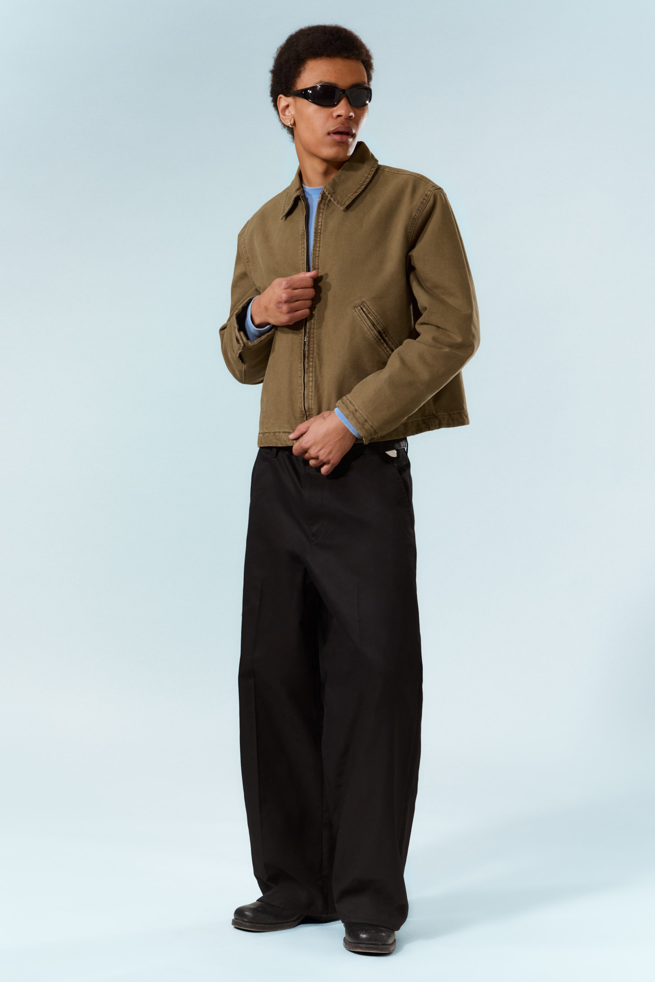 View larger image: An olive green work jacket is showcased in a three-quarter view, featuring a pointed collar, a central zip fastening, and two front patch pockets. It is layered over a light blue long-sleeved top, with black wide-leg trousers completing the outfit, exhibiting a loose fit.