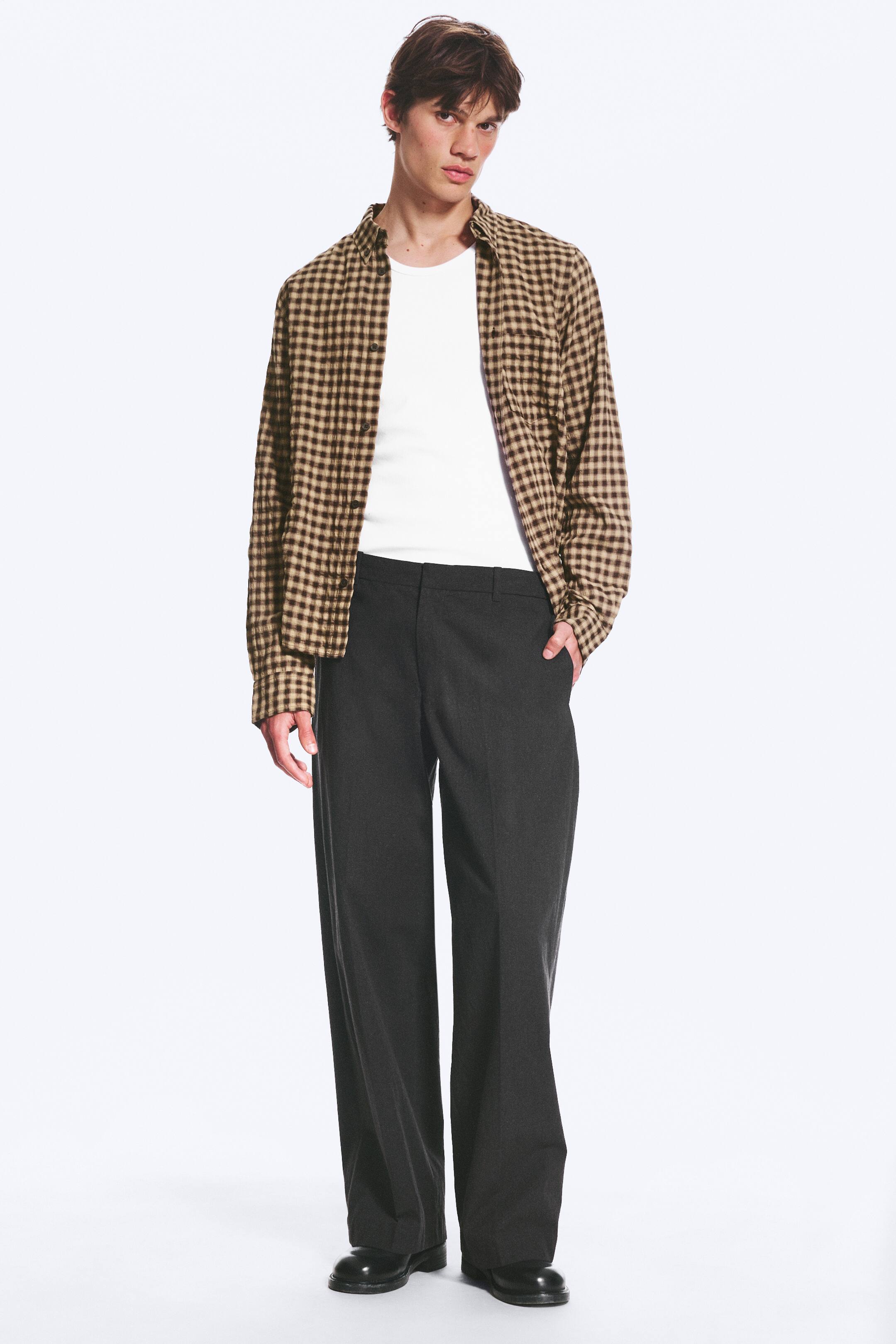Agrandir l'image: Une chemise à carreaux marron et beige à manches longues est portée ouverte sur un sous-vêtement à col rond blanc, associée à un pantalon large gris foncé et des chaussures noires.