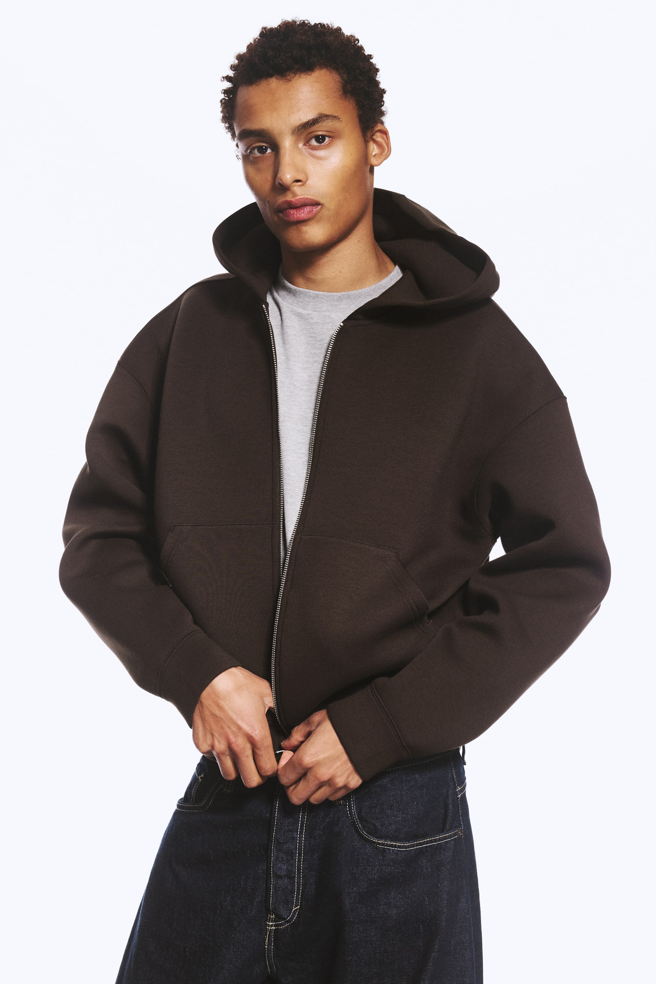 View larger image: A dark brown zip-up hoodie made from a thick scuba fabric is being worn by the model, who is adjusting the silver zip at the hem. The hoodie, featuring a spacious hood and dropped shoulders, is layered over a grey T-shirt and paired with dark blue jeans.