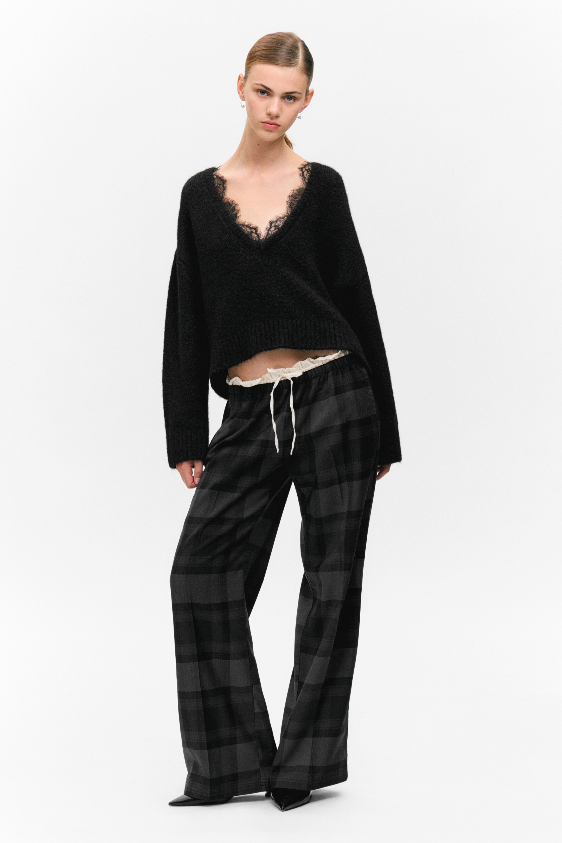Visualizza immagine più grande: Un maglione nero a V in maglia pelosa, con rifiniture in pizzo nero, mostra una vestibilità corta e rilassata sopra una canotta bianca con rifiniture in pizzo. Pantaloni a quadri grigio scuro e neri a gamba larga con un visibile cordoncino bianco sono anch'essi indossati.
