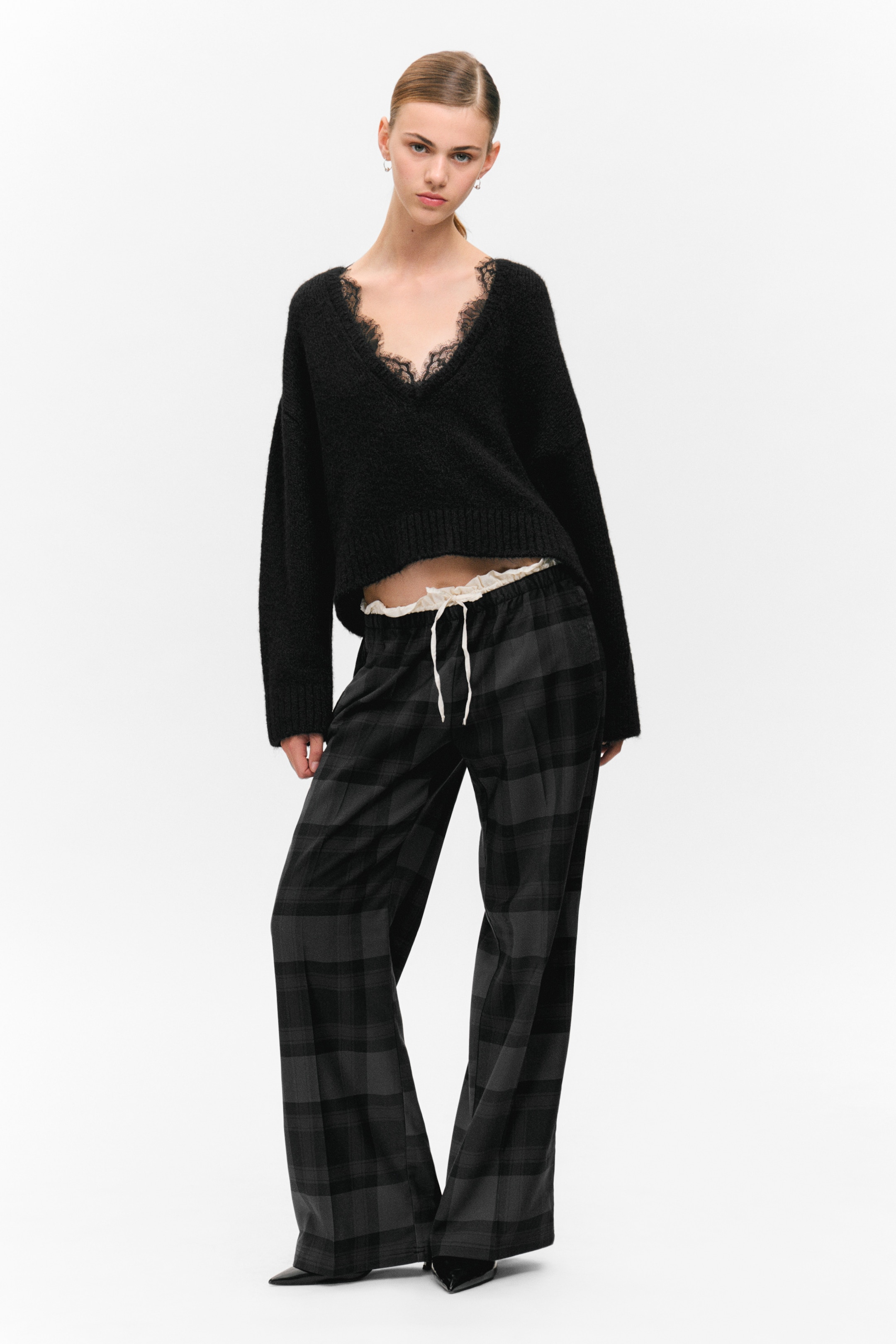 Ingrandisci l'immagine: Un maglione nero a V in maglia pelosa, con rifiniture in pizzo nero, mostra una vestibilità corta e rilassata sopra una canotta bianca con rifiniture in pizzo. Pantaloni a gamba larga a quadri grigio scuro e nero con un visibile cordoncino bianco sono anch'essi indossati.