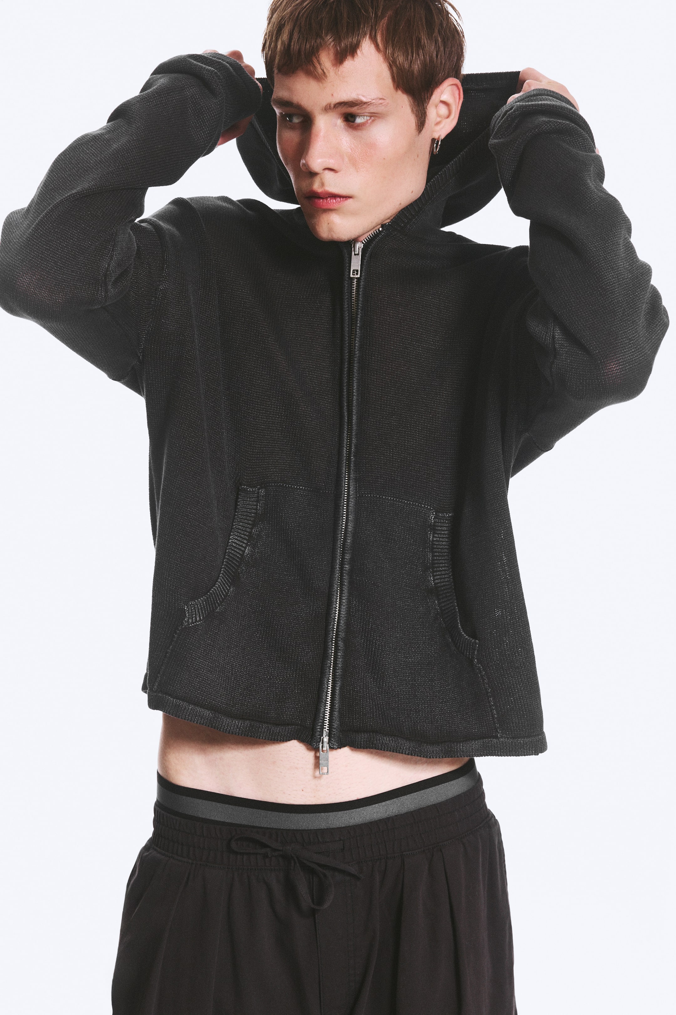 Grotere afbeelding bekijken: Een donker getextureerde gebreide hoodie met volledige ritssluiting wordt gedragen door het model, die de capuchon aanpast. De korte top toont een gedeeltelijk geopende metalen voorrits en twee opgestikte zakken, en is gestyled over donkere shorts met een elastische tailleband.