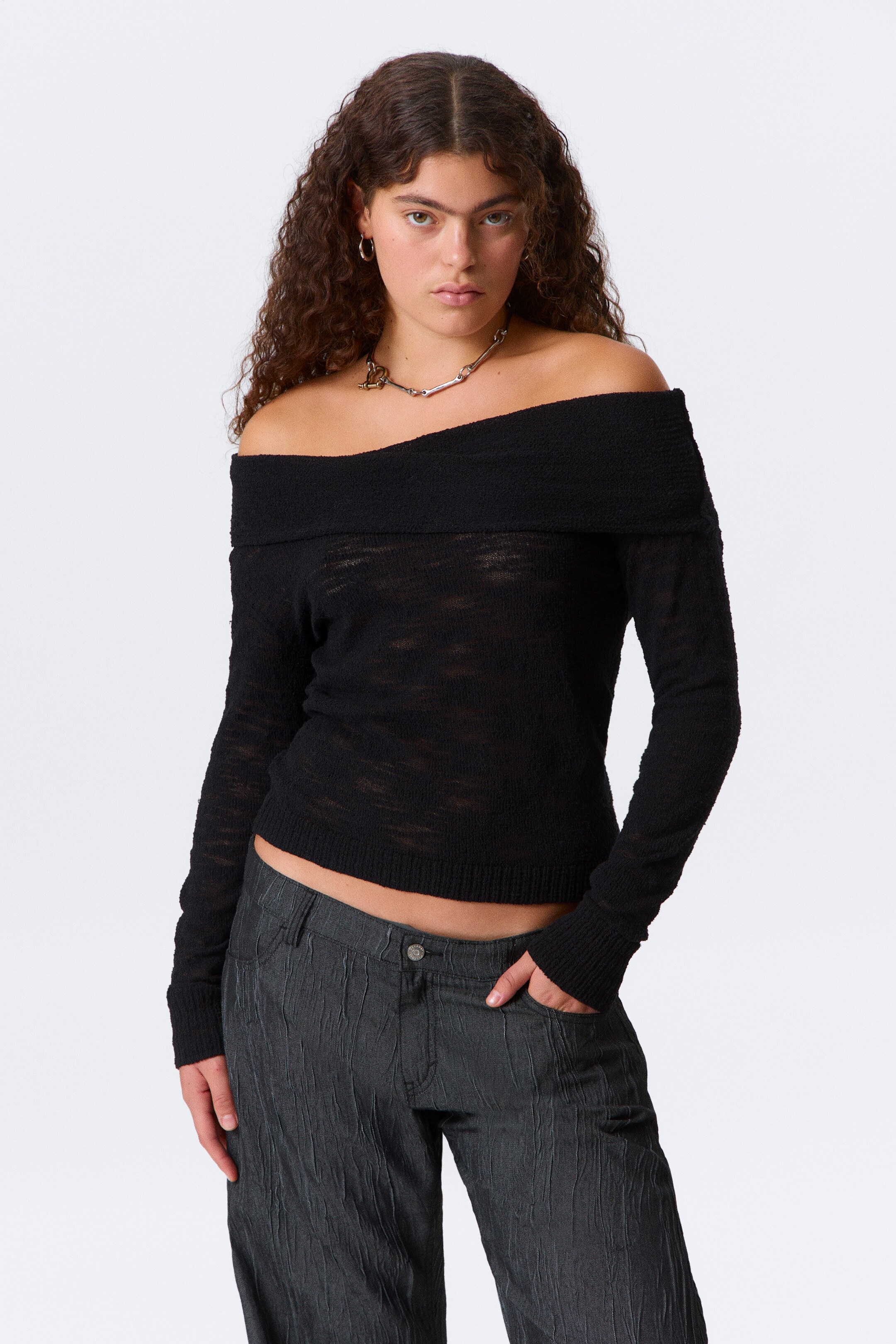 Visa större bild: En svart stickad off-shoulder-tröja bärs av modellen. Tröjan har en bred vikt halsringning, en transparent öppen stickad struktur och en beskuren, något figurnära siluett med ribbstickade muddar och fåll. Ett halsband i silver och guldfärgad ton vilar på tröjans halsringning.