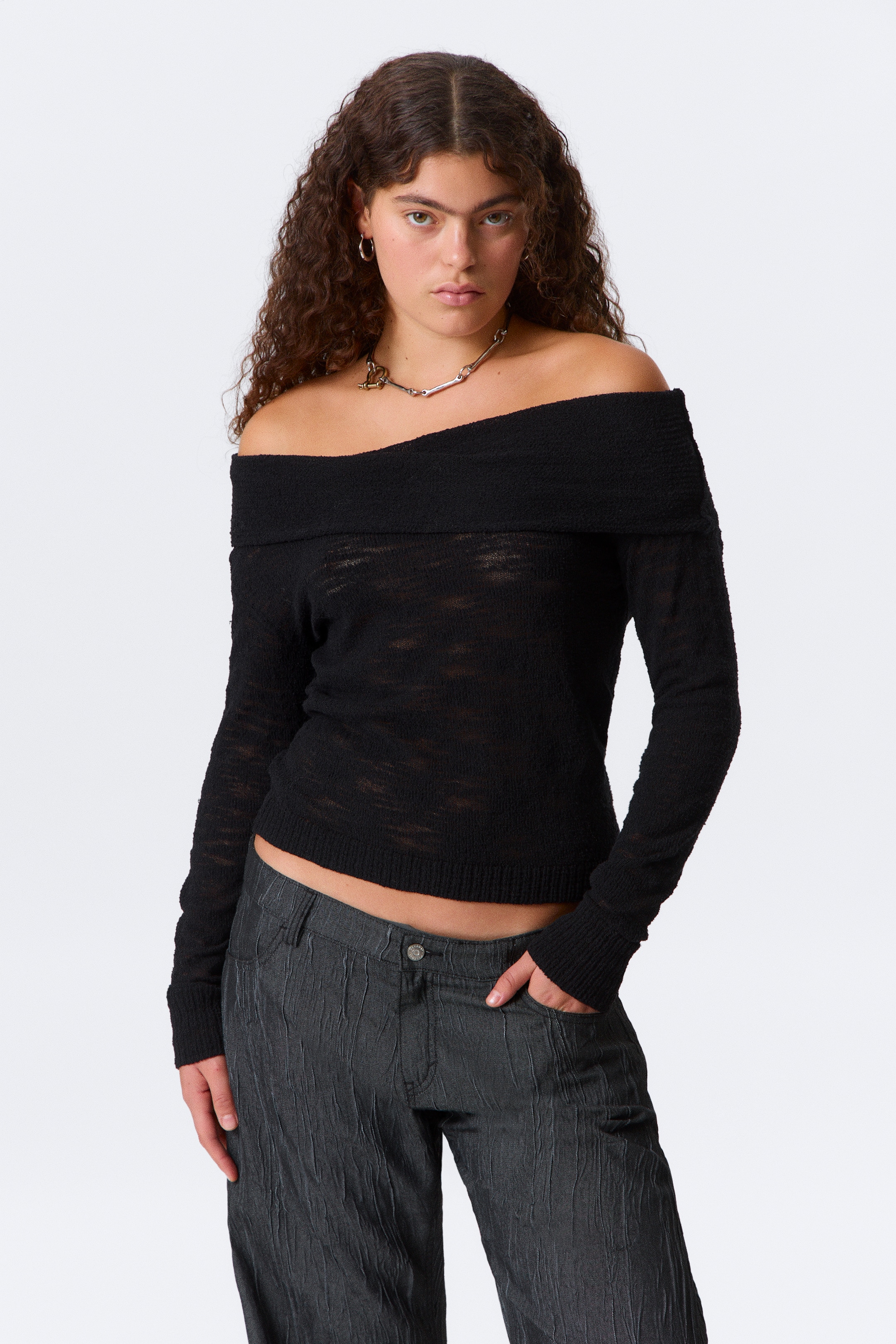 Visualizza immagine più grande: Un maglione nero a spalle scoperte lavorato a maglia è indossato dalla modella. Il maglione presenta un ampio scollo ripiegato, una texture in maglia aperta trasparente e una silhouette corta, leggermente aderente, con polsini e orlo a coste. Una collana dai toni argento e oro è appoggiata sullo scollo del maglione.