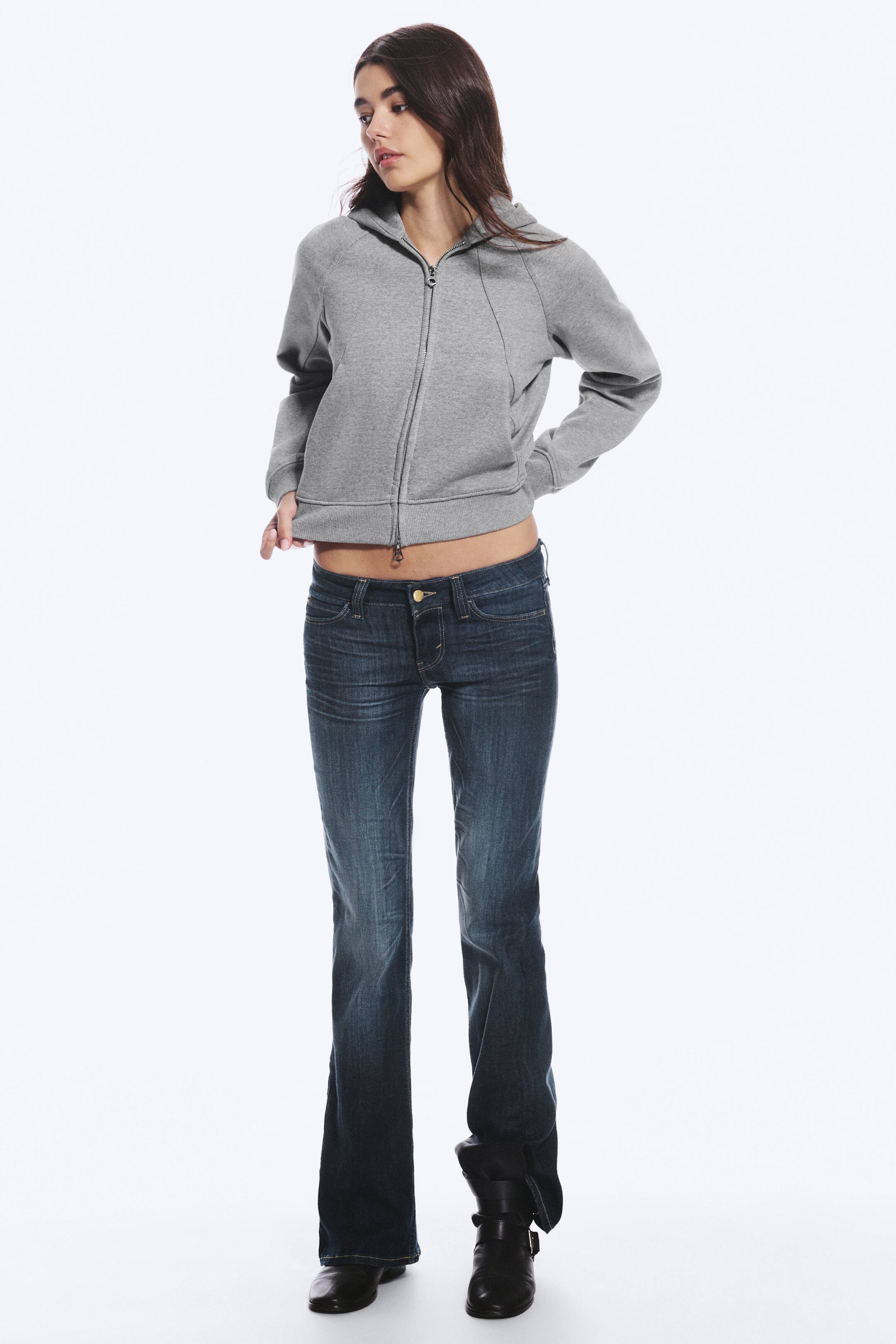 View larger image: Low-rise, dark-wash bootcut jeans with subtle fading on the thighs are being worn by the model, who is lifting the hem of a grey zip-up hoodie to reveal the low waistband. The jeans flare slightly at the bottom and are paired with black boots.