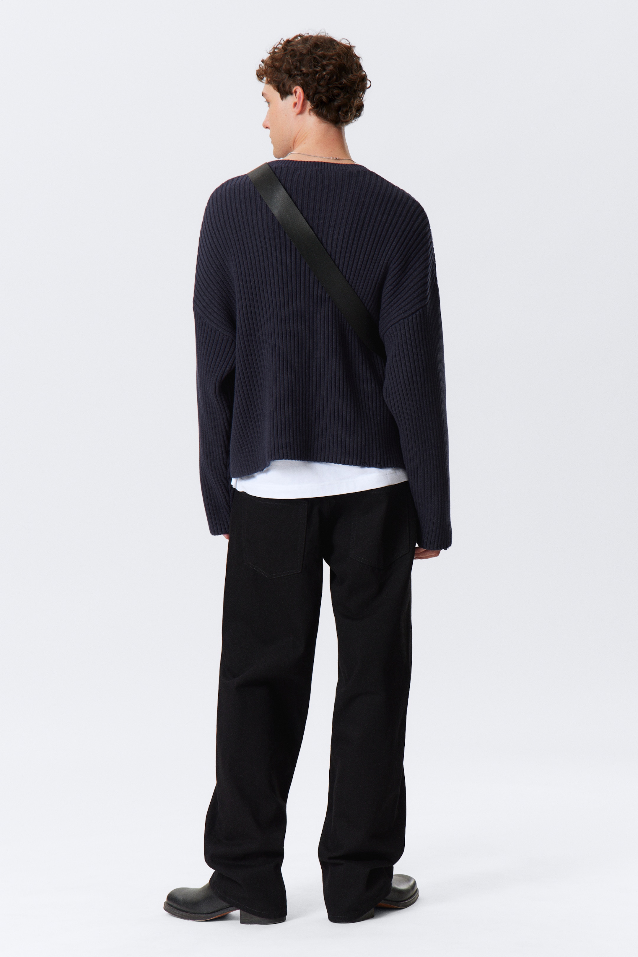 Visualizza immagine più grande: Un maglione a coste blu navy viene presentato dal retro, mettendo in mostra la sua vestibilità rilassata, le spalle cadenti e l'orlo grezzo e corto. Una t-shirt bianca è visibilmente stratificata sotto il maglione, mentre una cinghia nera lo attraversa. Sono visibili anche pantaloni neri a gamba larga con tasche posteriori applicate, che si appoggiano su scarpe nere massicce.